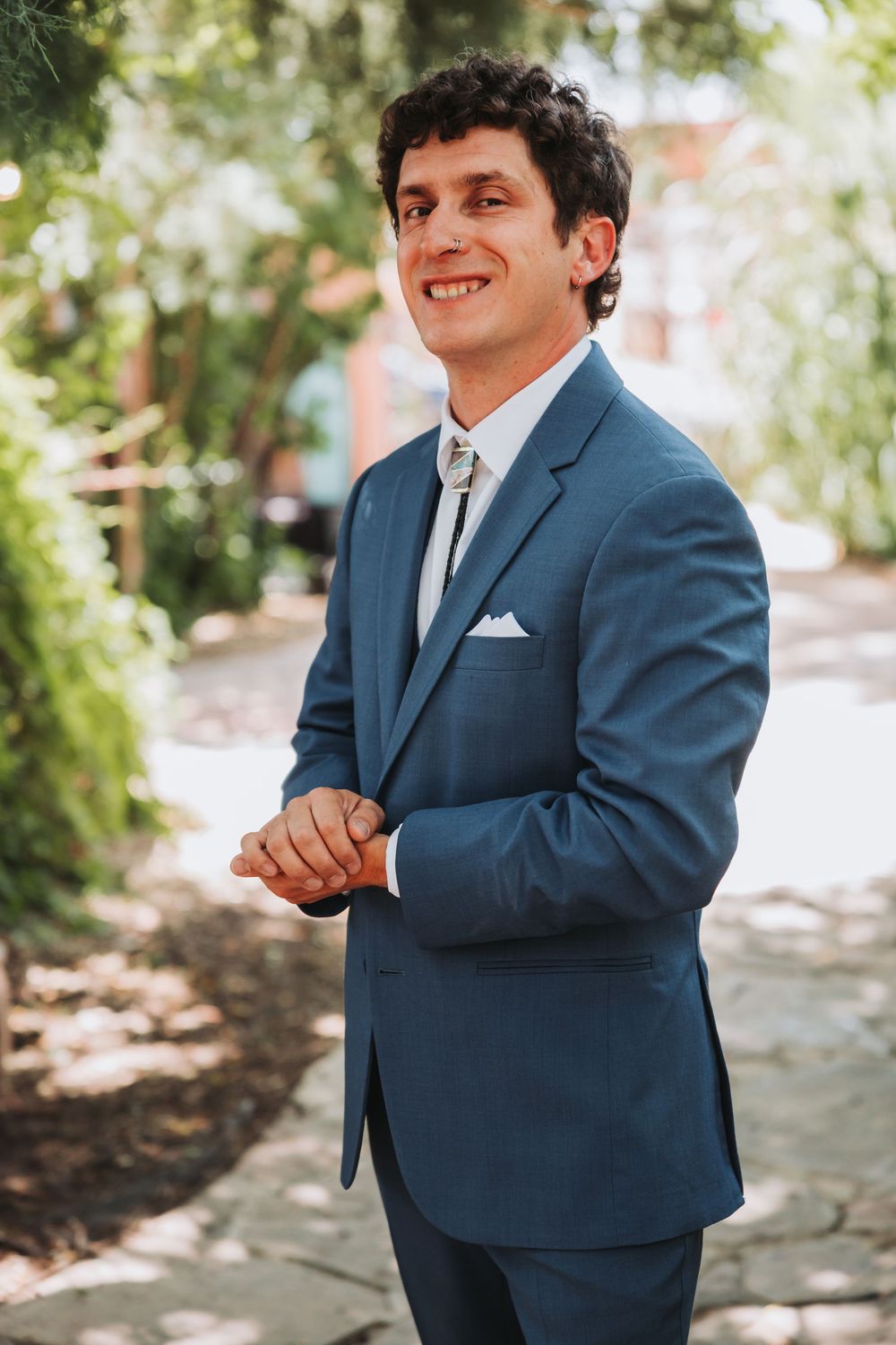 A person wearing a classic blue suit with white pocket square stands in a garden setting with dappled sunlight.