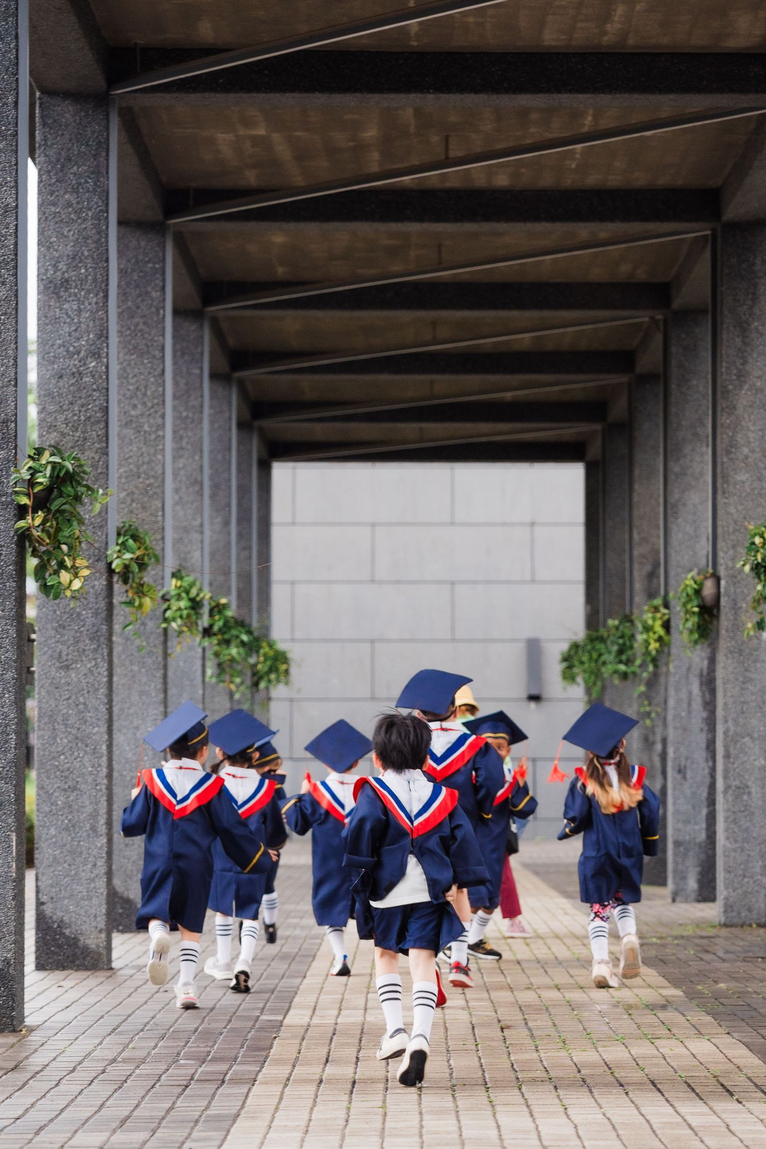 Young graduates in navy blue and red uniforms with mortarboards running through a covered walkway decorated with greenery.