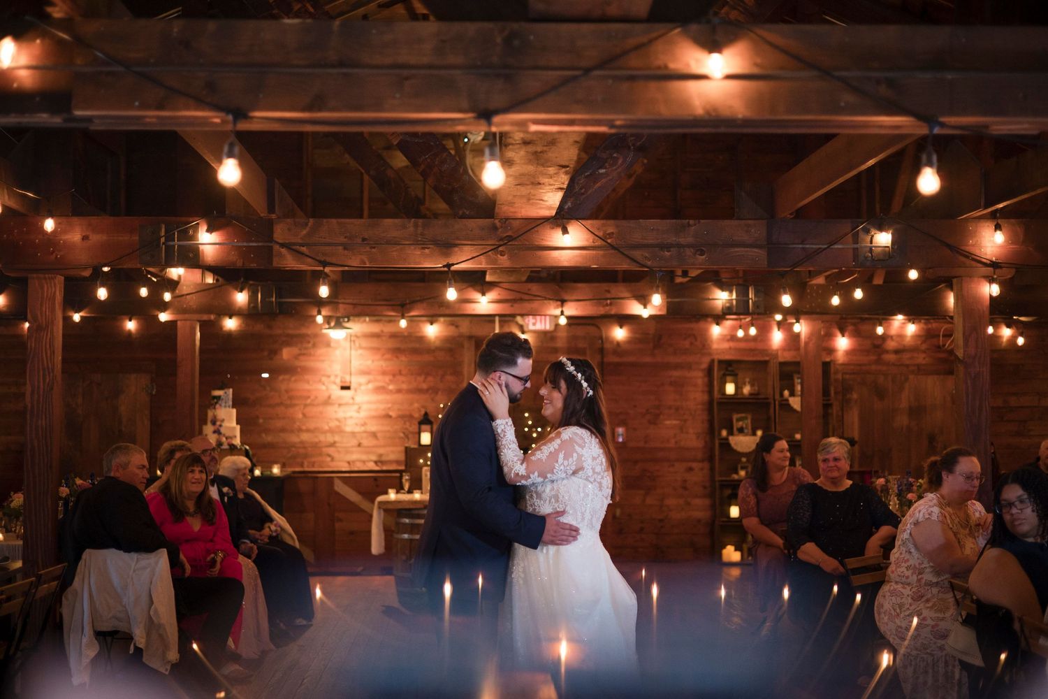 Wedding reception in a rustic barn venue illuminated by warm string lights and glowing candles along wooden tables.