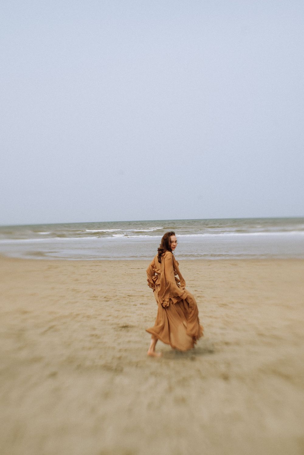 A brown dress flows gracefully across a misty beach landscape.