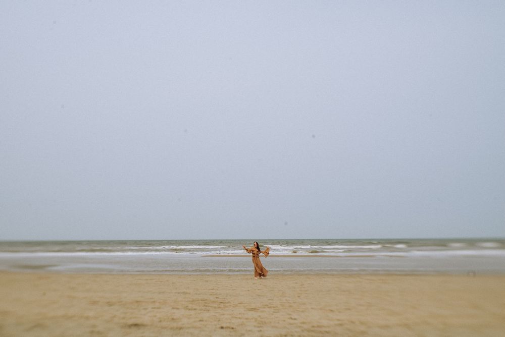 A figure in flowing fabric dances on an empty beach with waves in the background.