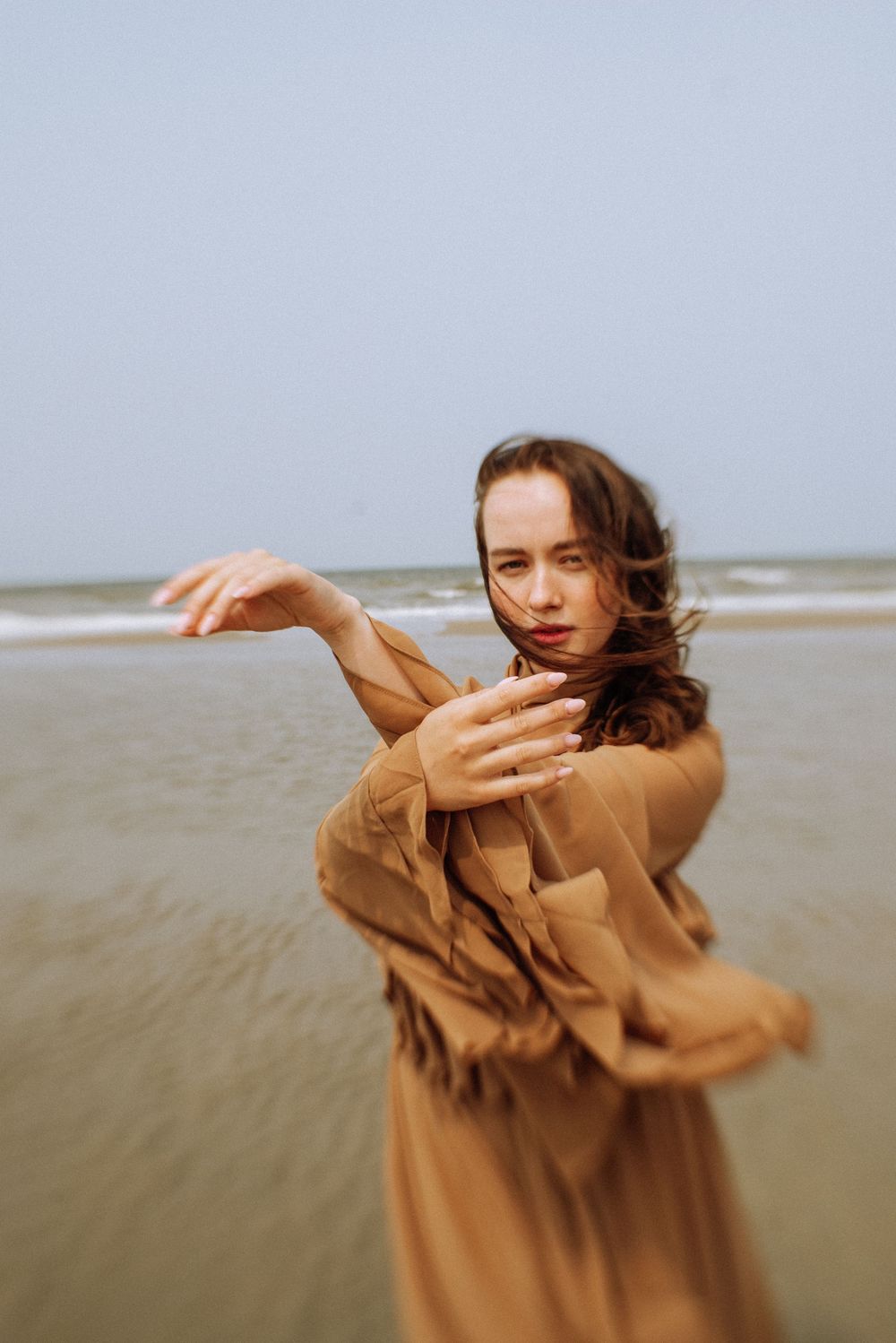 A dancer in brown fabric moves expressively on a windswept beach.