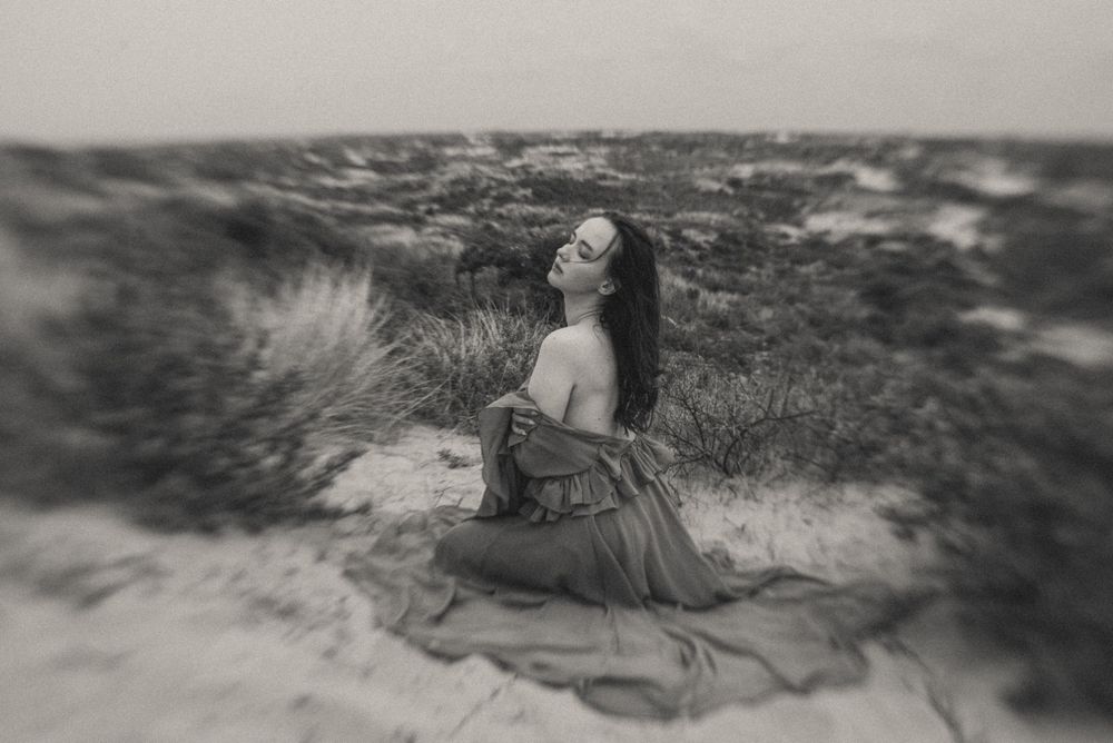 A dreamy black and white portrait shows a figure reclining in coastal dunes.