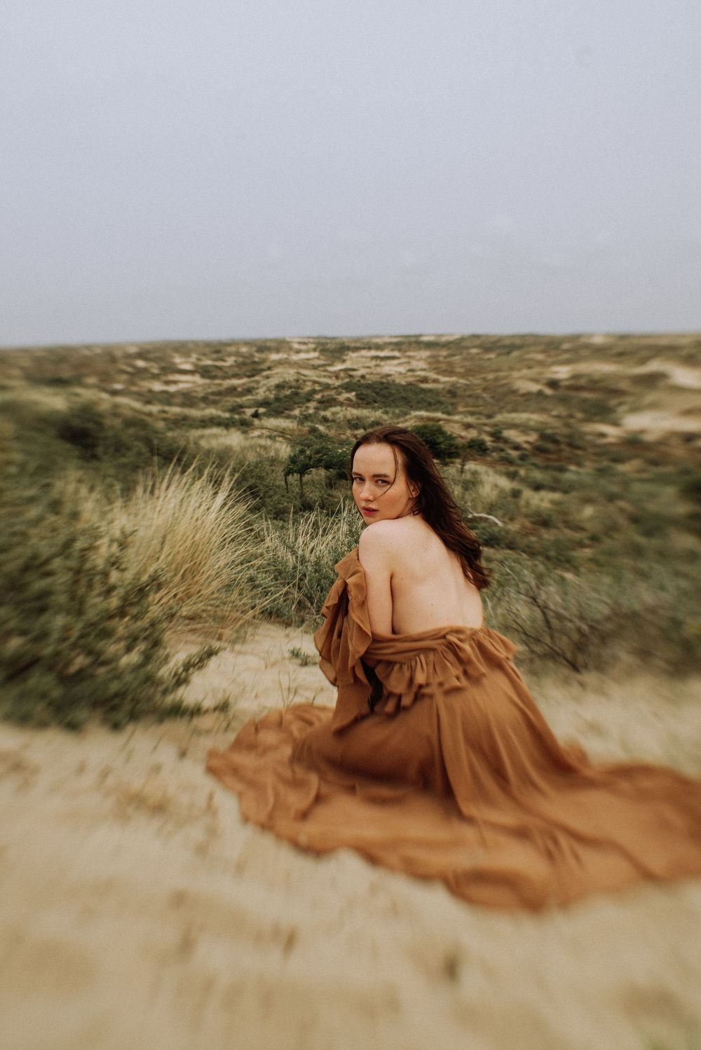 A brown dress flows across coastal dunes with wild grasses in soft focus.