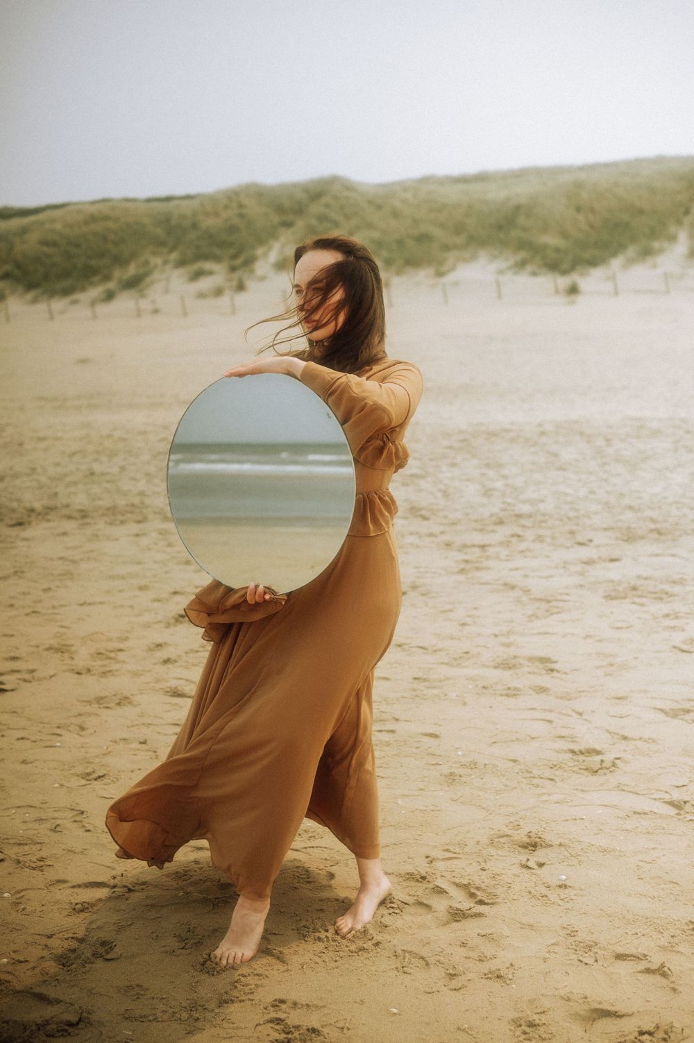 A person in a brown dress moves expressively across sandy beach dunes at sunset.