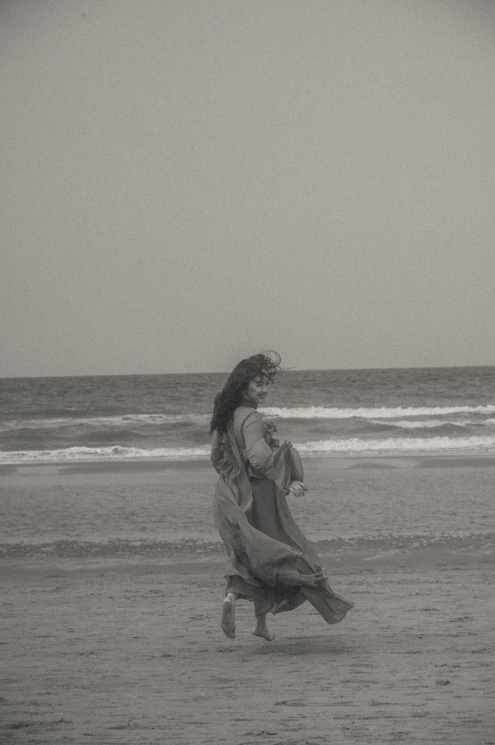A figure in a flowing dress dances gracefully along a misty beach shoreline in black and white.