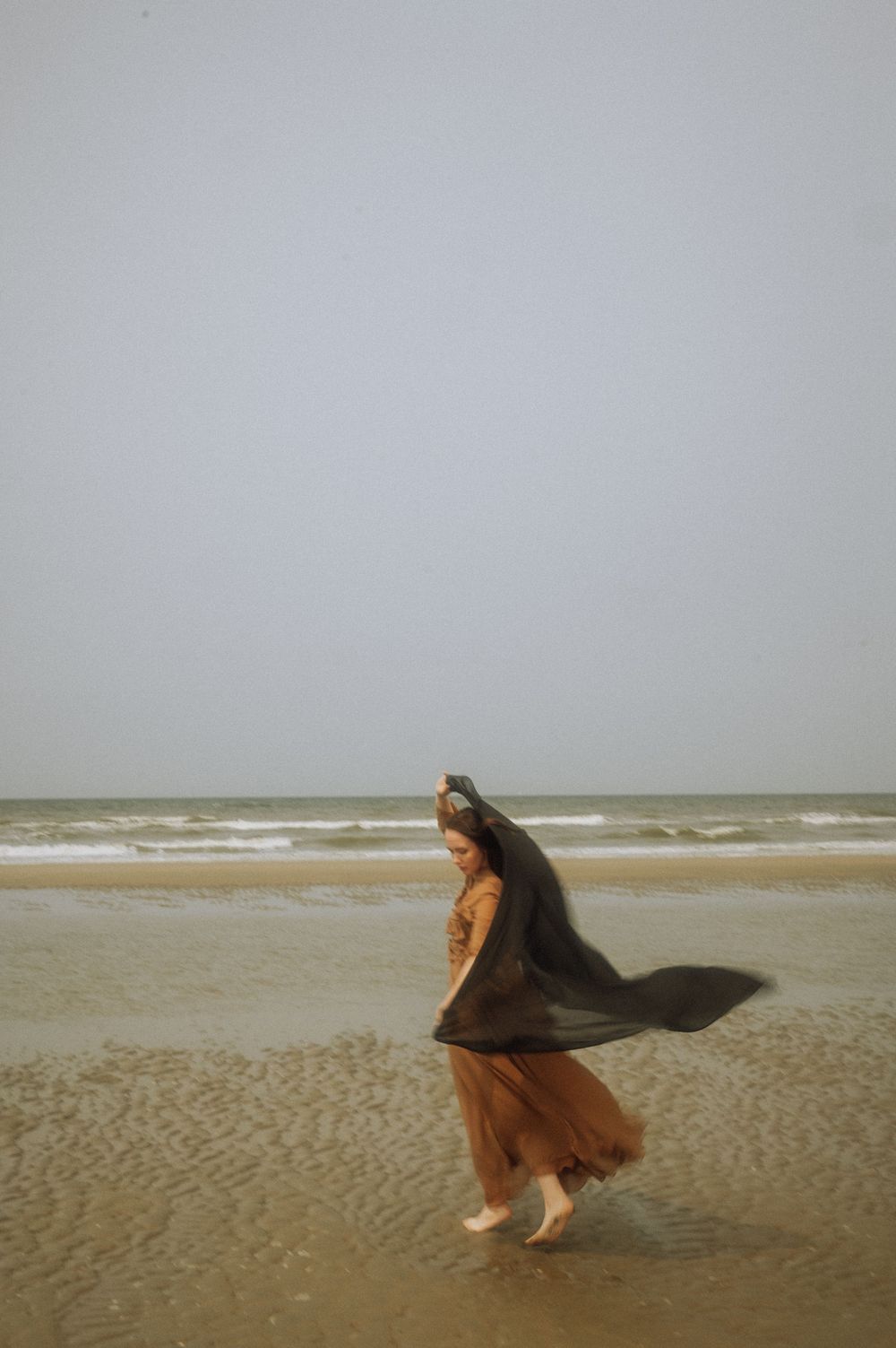 A flowing brown dress catches the coastal wind on an overcast beach.