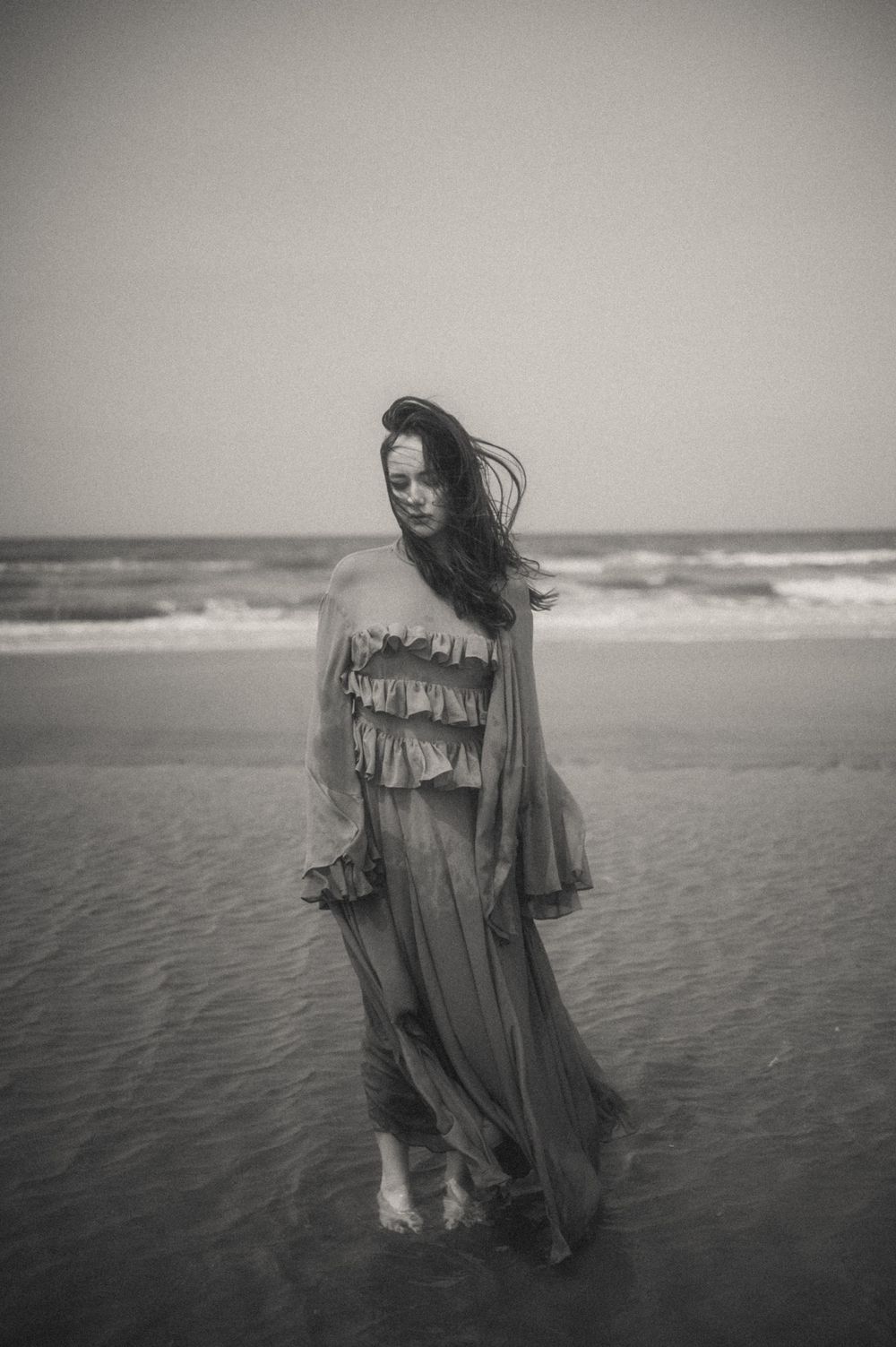 An ethereal figure in a flowing dress stands in shallow ocean waves in black and white.