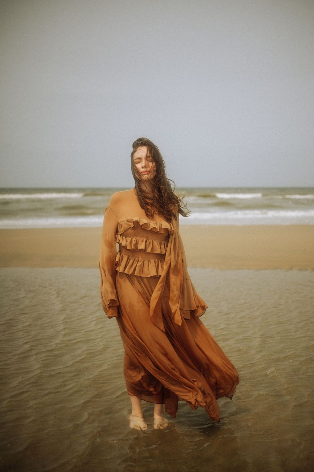 A flowing brown dress billows in the ocean breeze as waves lap at bare feet on the shore.