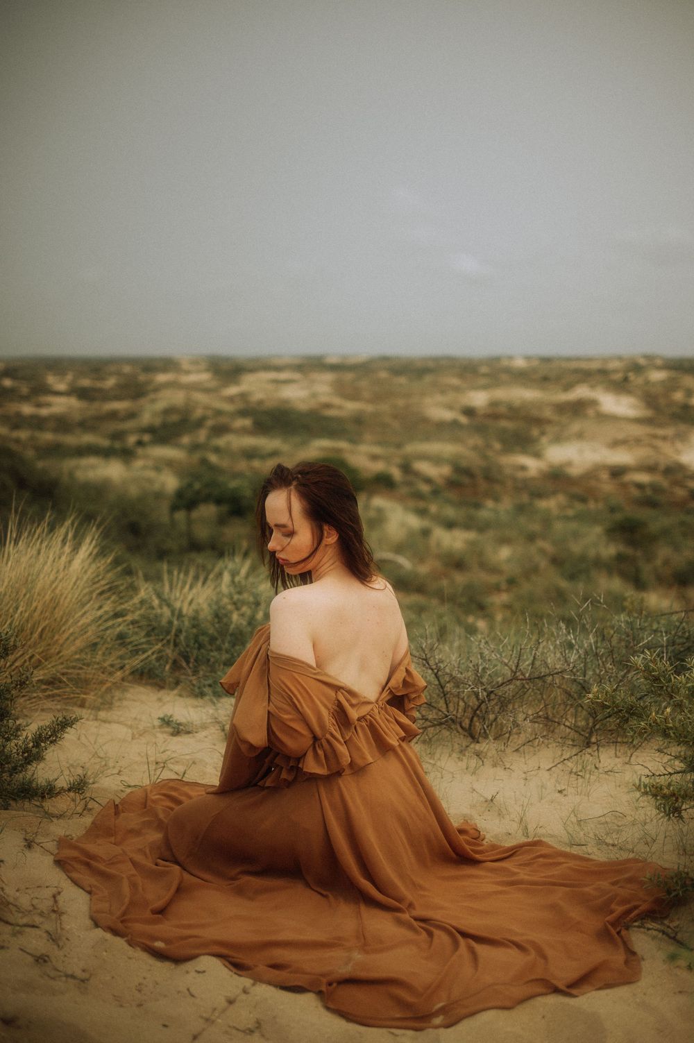 A flowing brown dress billows dramatically against desert sand dunes and sparse vegetation.