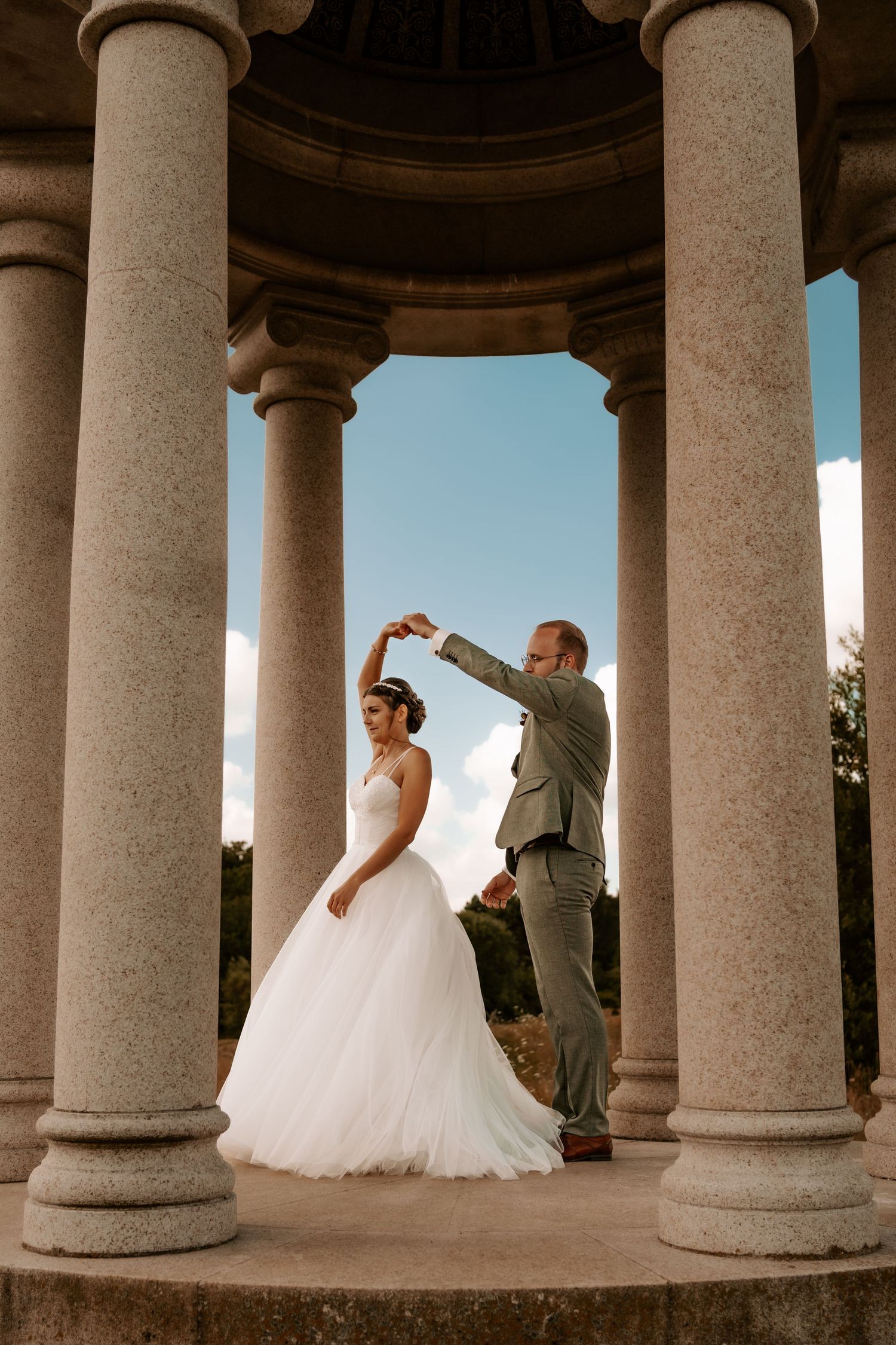Wedding couple dances between elegant stone columns in a romantic outdoor venue at golden hour.