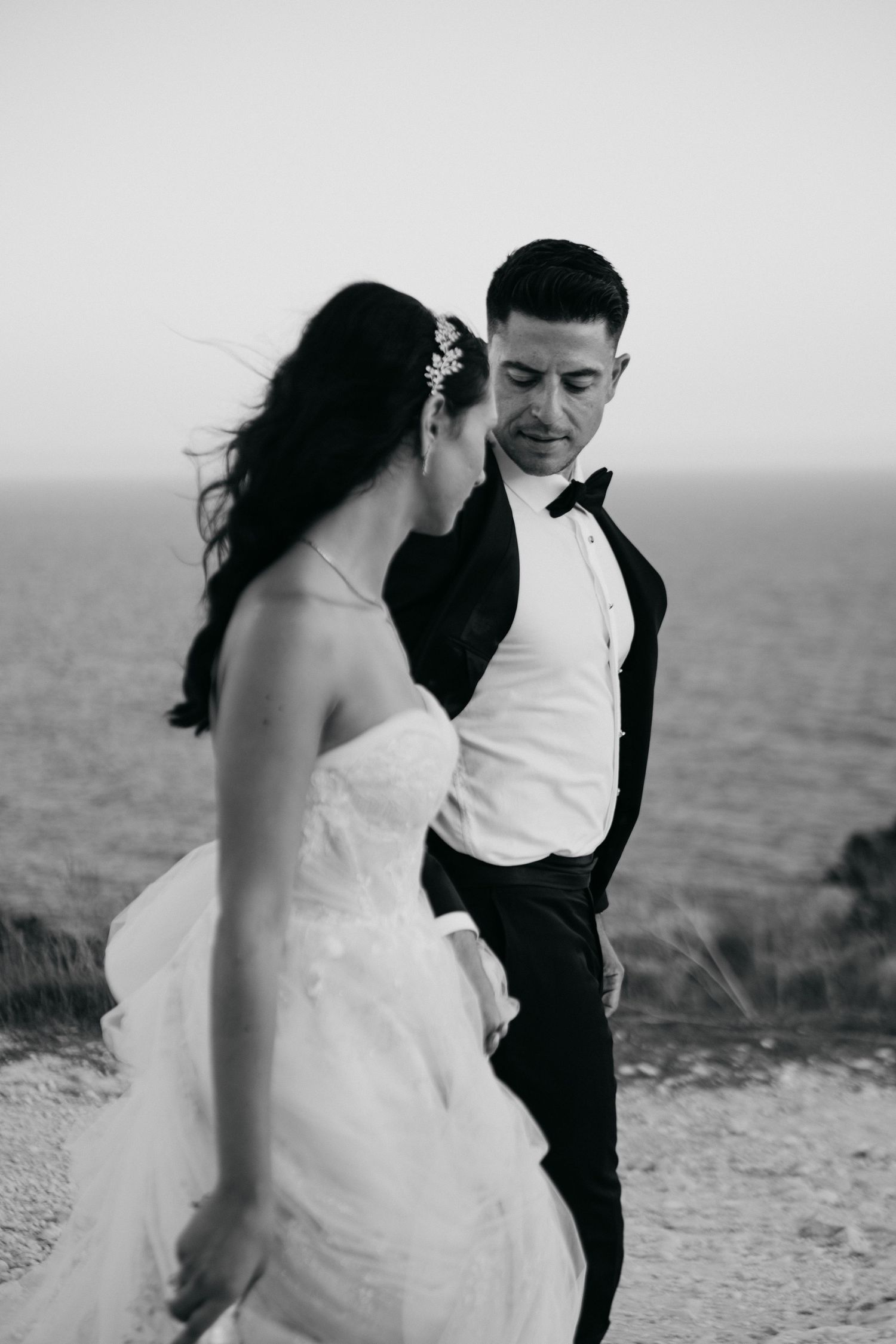 A bride in a flowing white wedding dress with lace back detail stands on a sandy dune in a desert landscape.