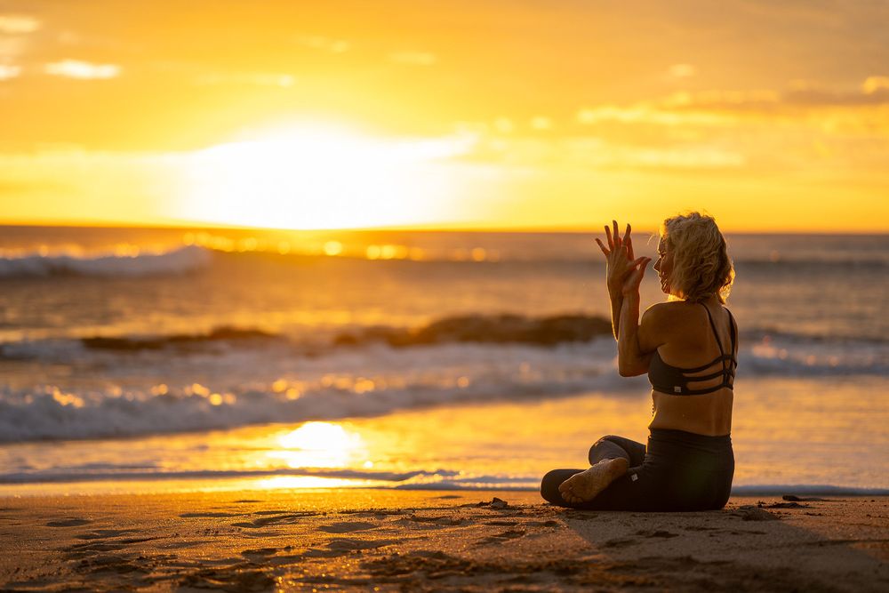 A silhouette of someone meditating on a beach at sunset with golden waves crashing in the background.