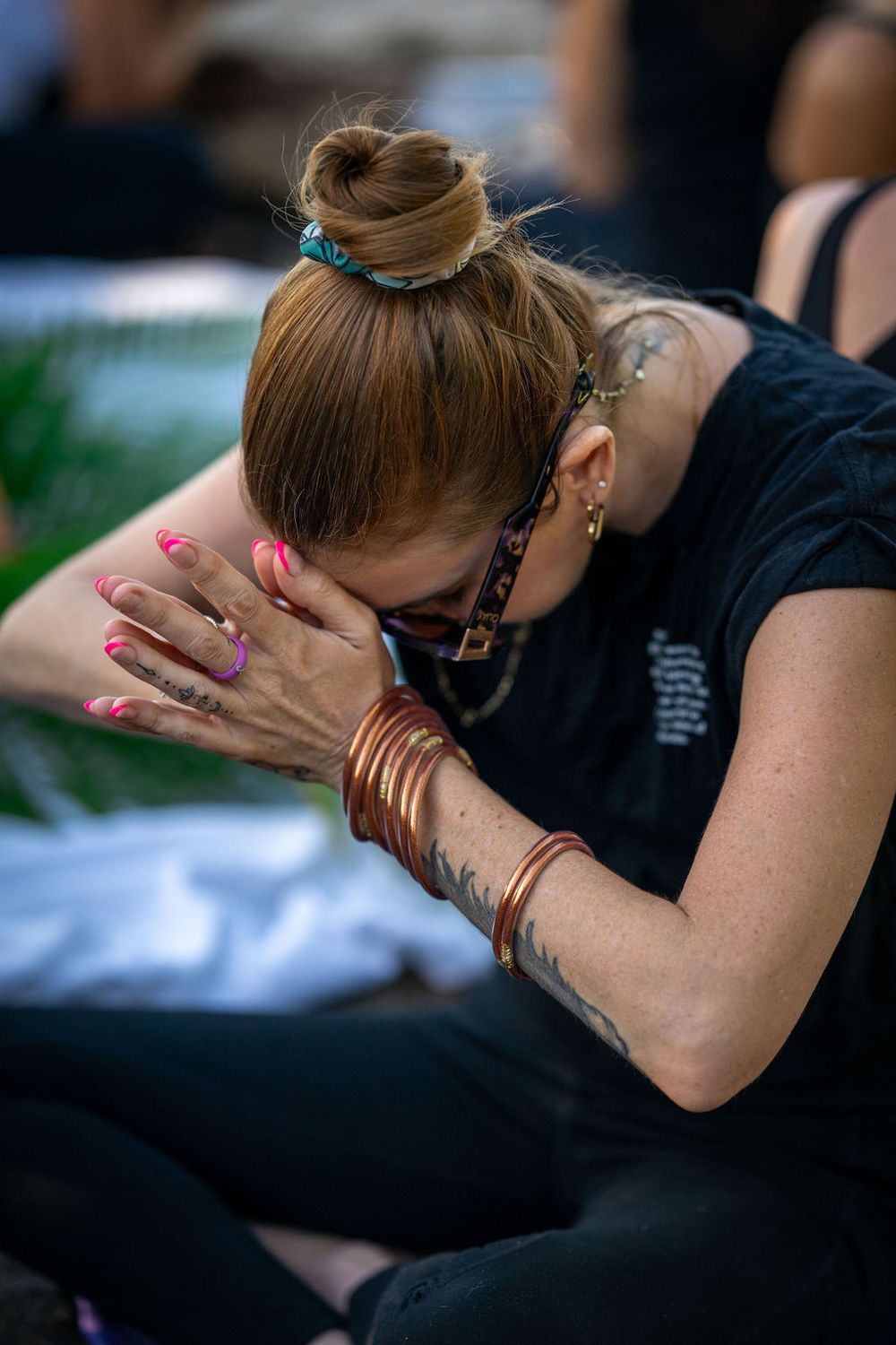 Someone with a bun hairstyle bows head in prayer while wearing bracelets and displaying pink nail polish.