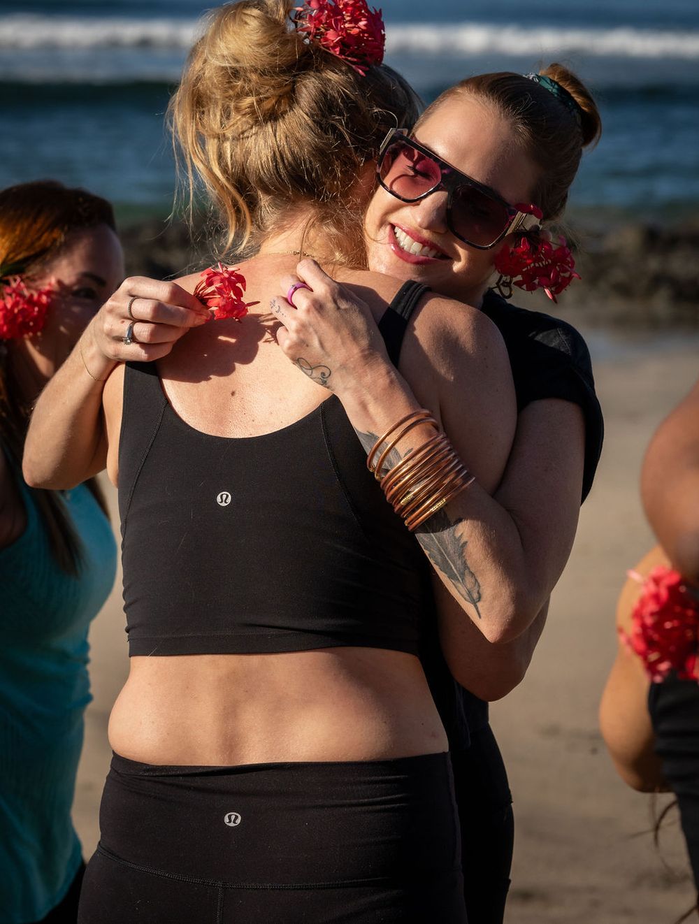 Friends in matching black workout attire share a joyful moment together on the beach with red flowers in their hair.