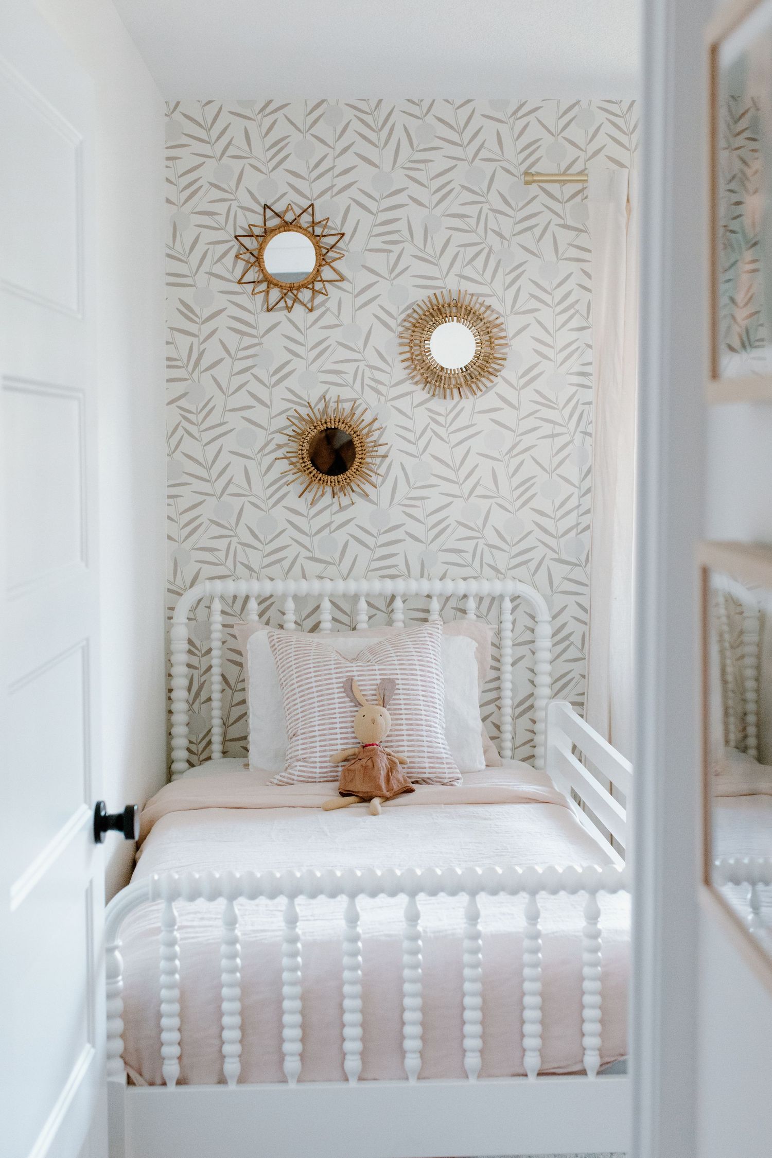 White bedroom with geometric wallpaper, spindle bed, and decorative sun mirrors on the wall.