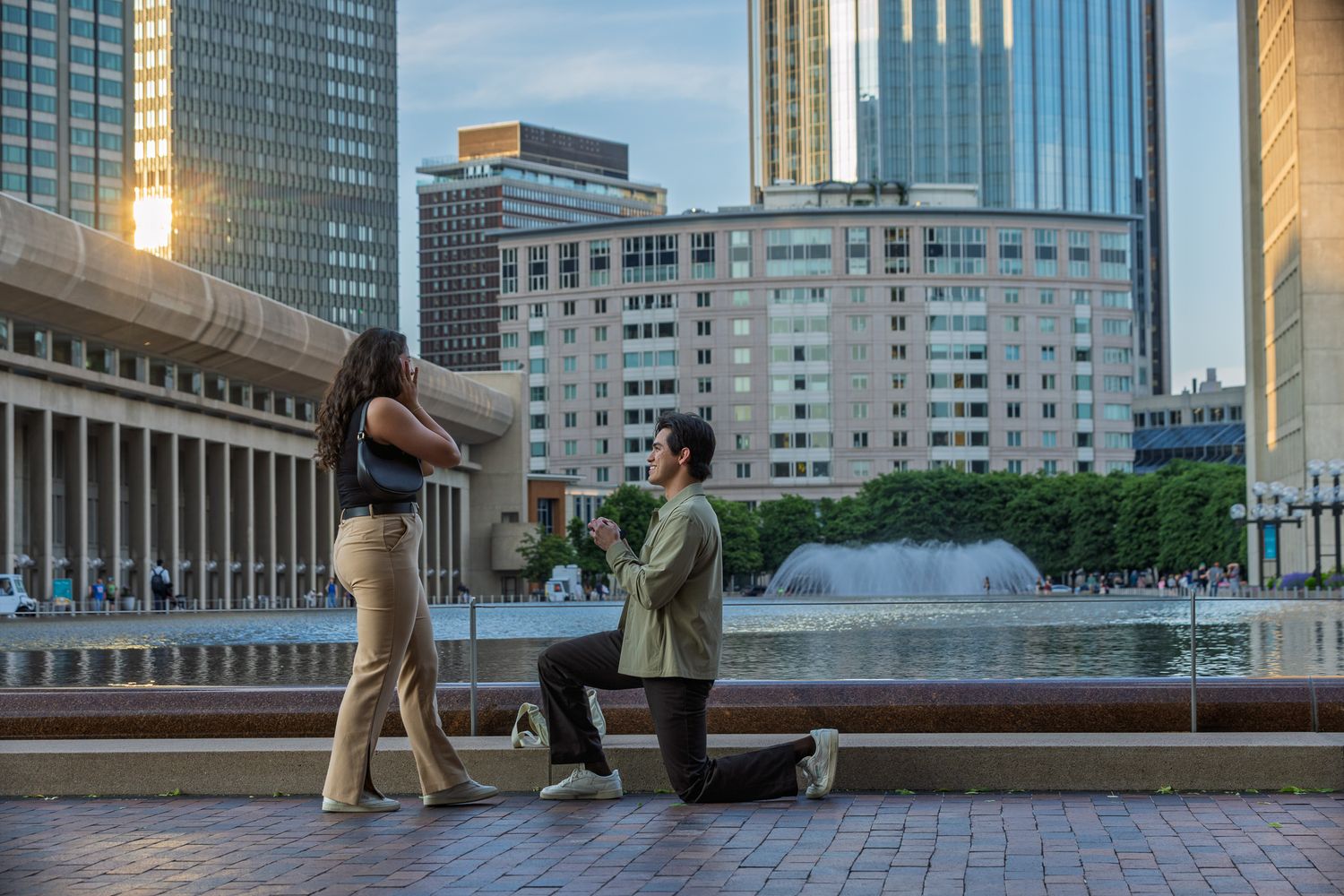 A romantic marriage proposal scene by a fountain with modern city buildings in the background during golden hour.