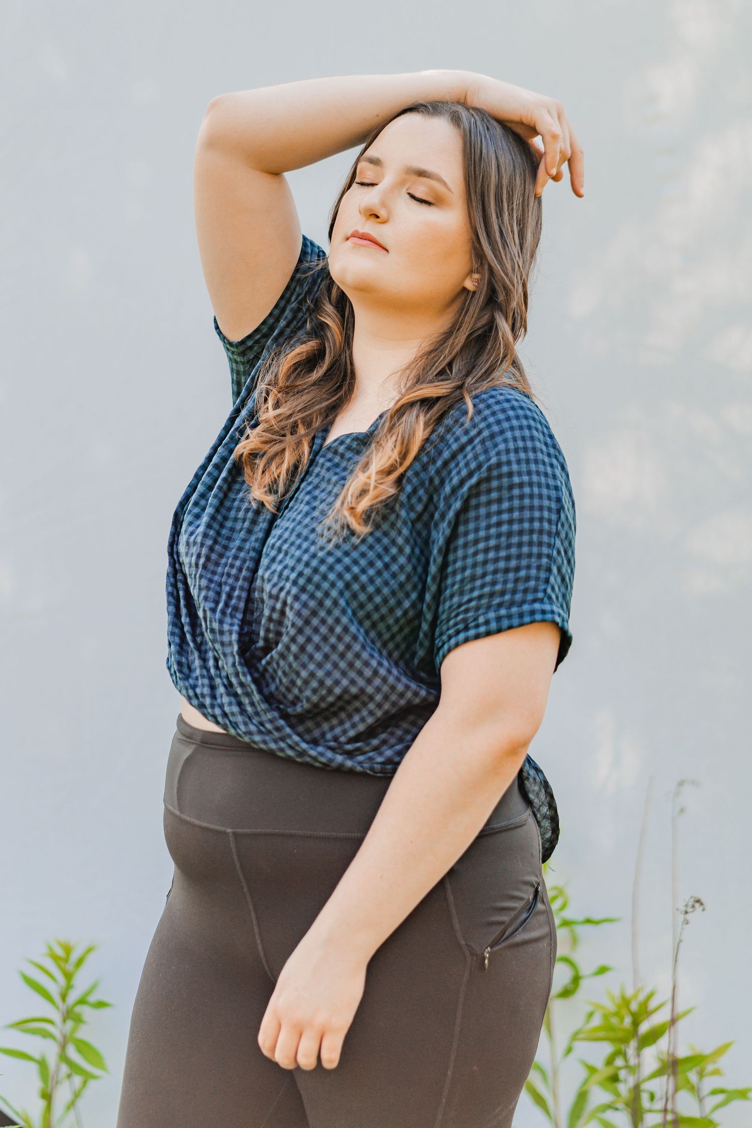 Portrait of a woman with eyes closed and hand in hair, standing against a neutral backdrop outdoors in natural light.