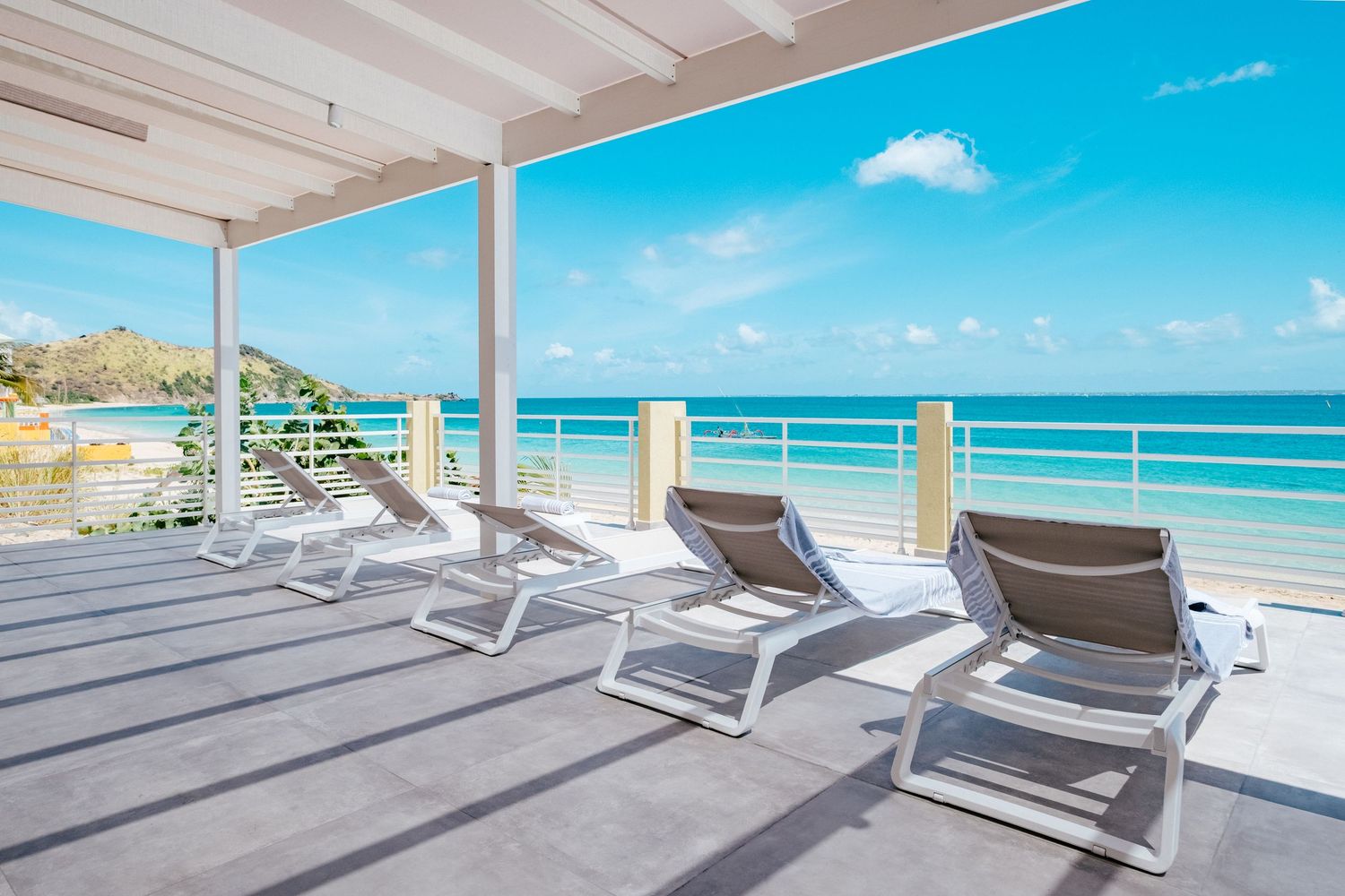 Luxury beachfront balcony with white lounge chairs overlooking turquoise Caribbean waters and blue sky.