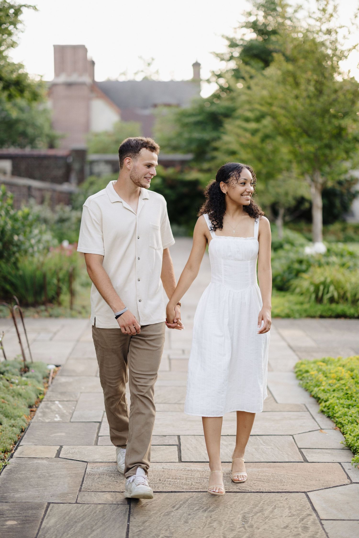 Couple walking hand in hand on stone path in garden, with person in white dress and khaki pants smiling together.