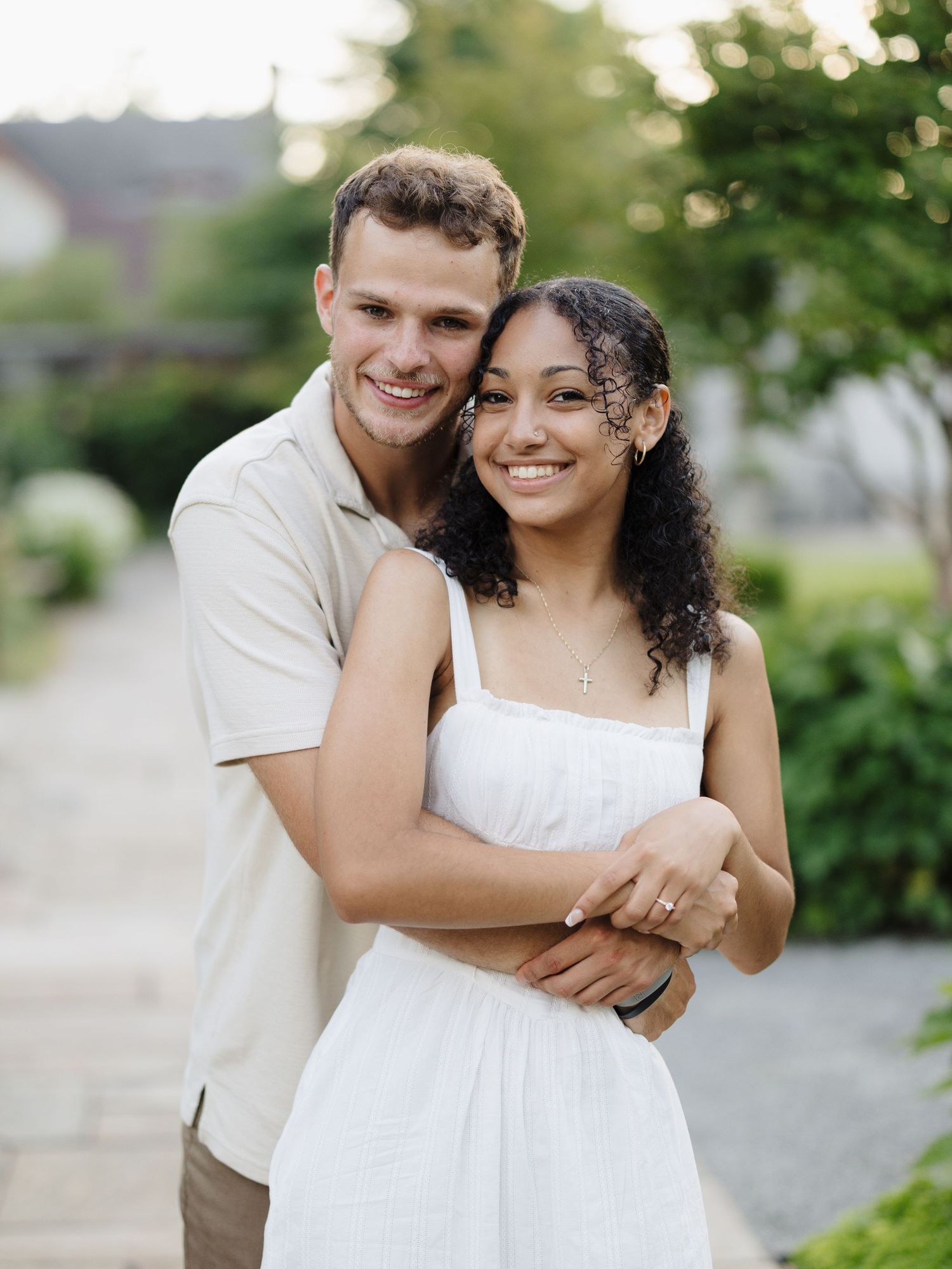 A couple shares a loving embrace outdoors on a sunny day, with lush greenery and bokeh background effect.