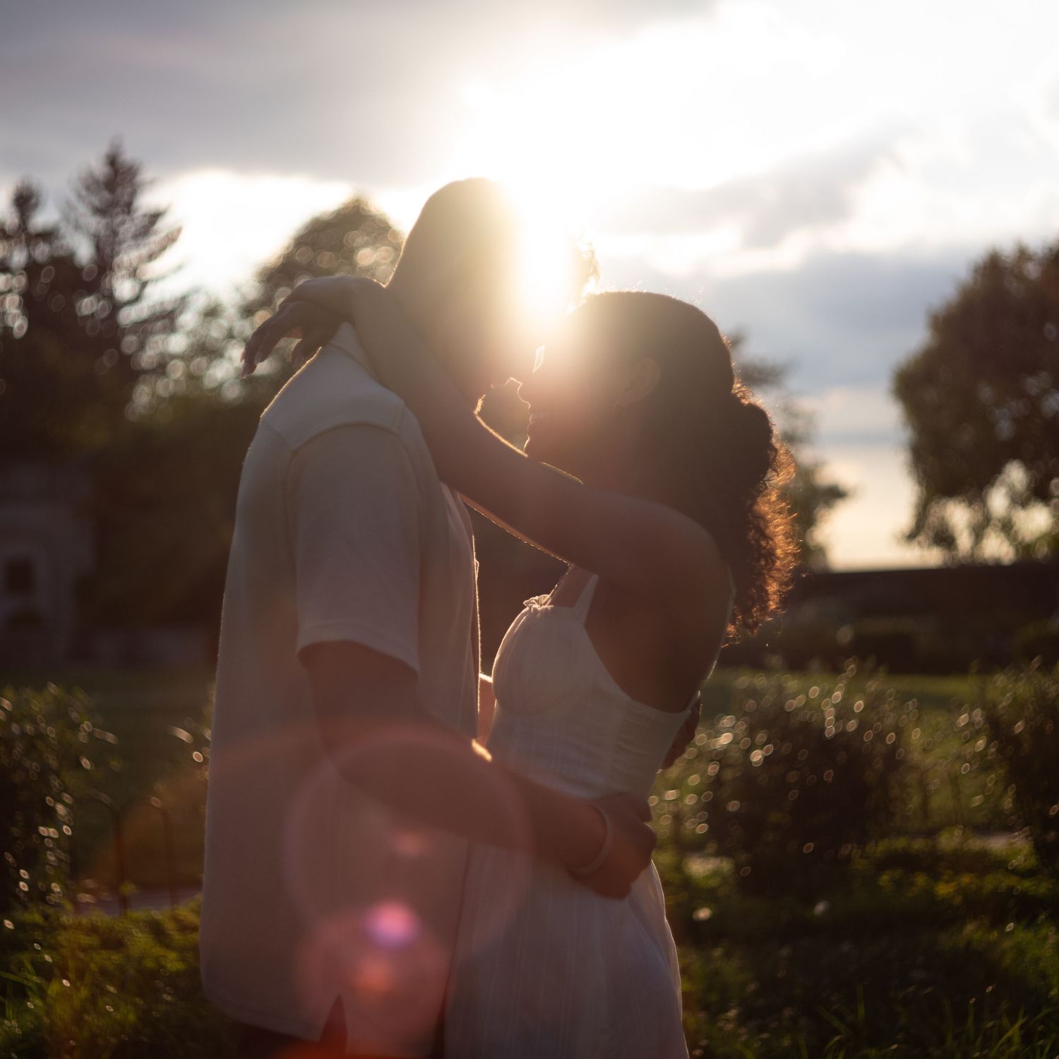 A couple embraces in silhouette against a golden sunset with sun flare and trees in the background.