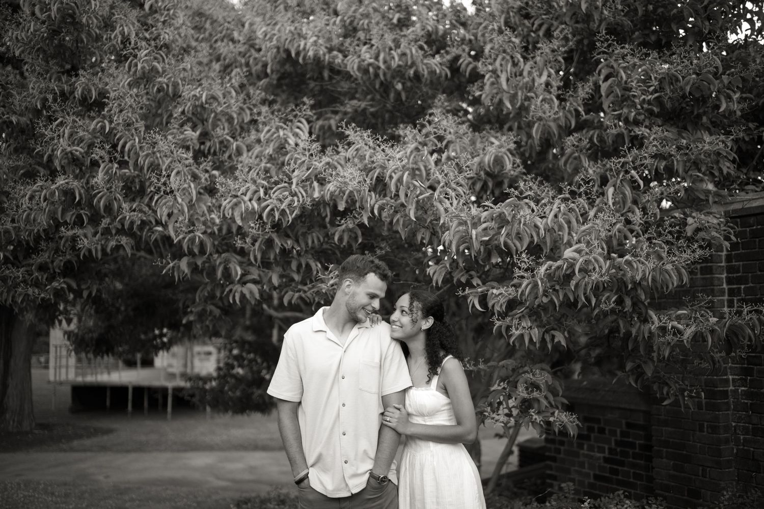 Romantic black and white portrait of couple standing together under leafy trees in garden setting.