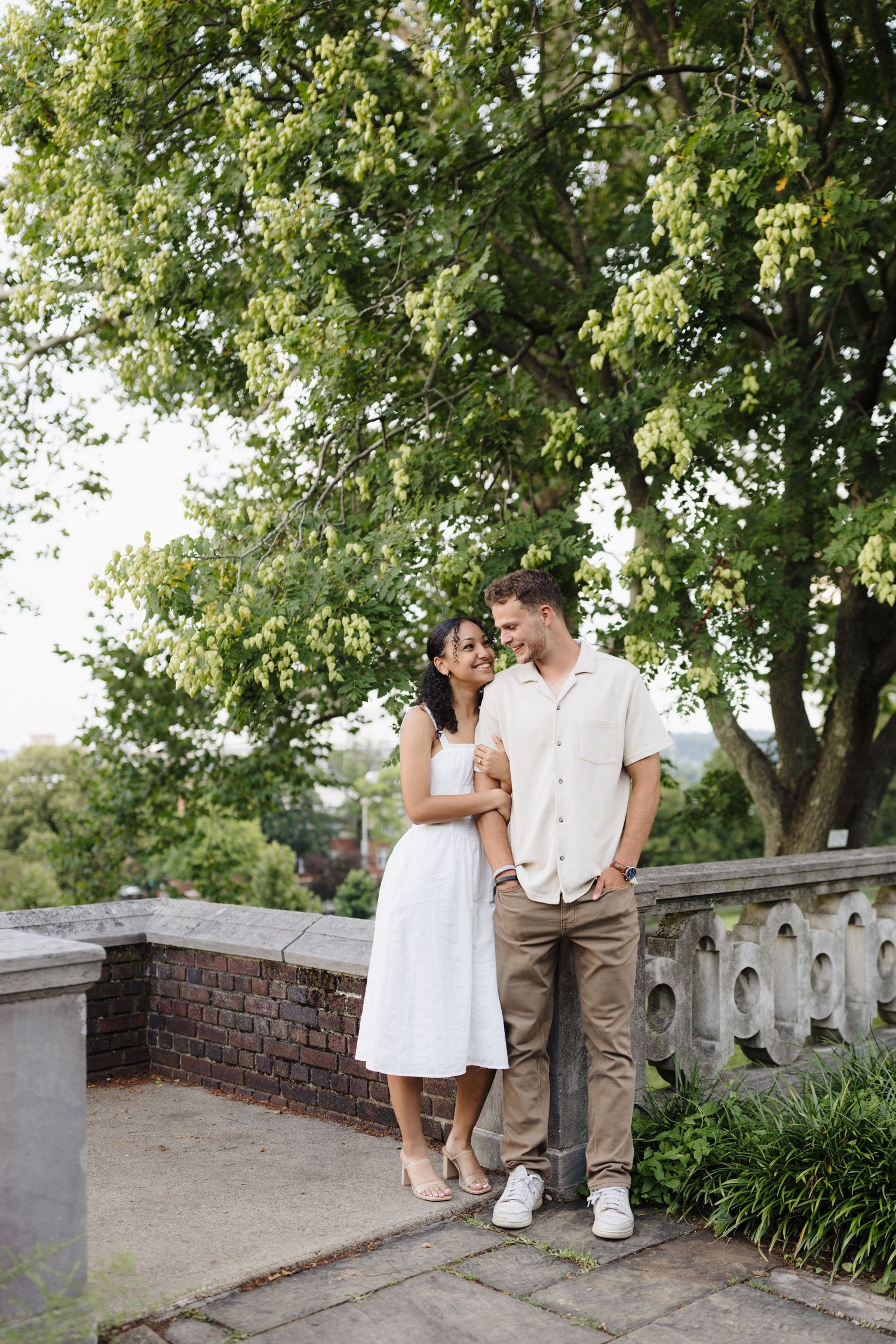 A couple shares tender moments together on a brick wall walkway surrounded by lush green trees on a summer day.