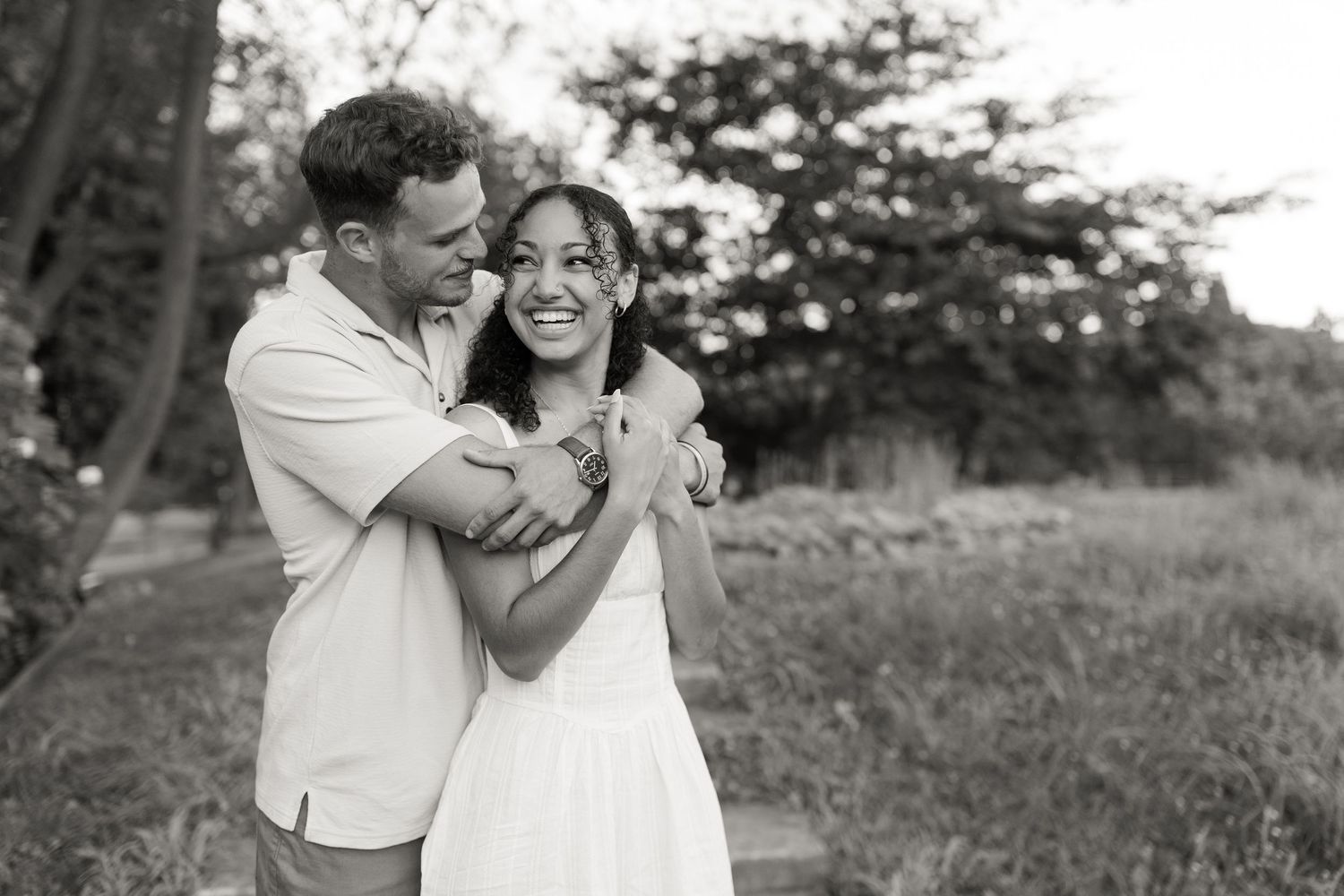 Couple in white outfits shares a joyful embrace while standing in a grassy outdoor setting during a sunny day.