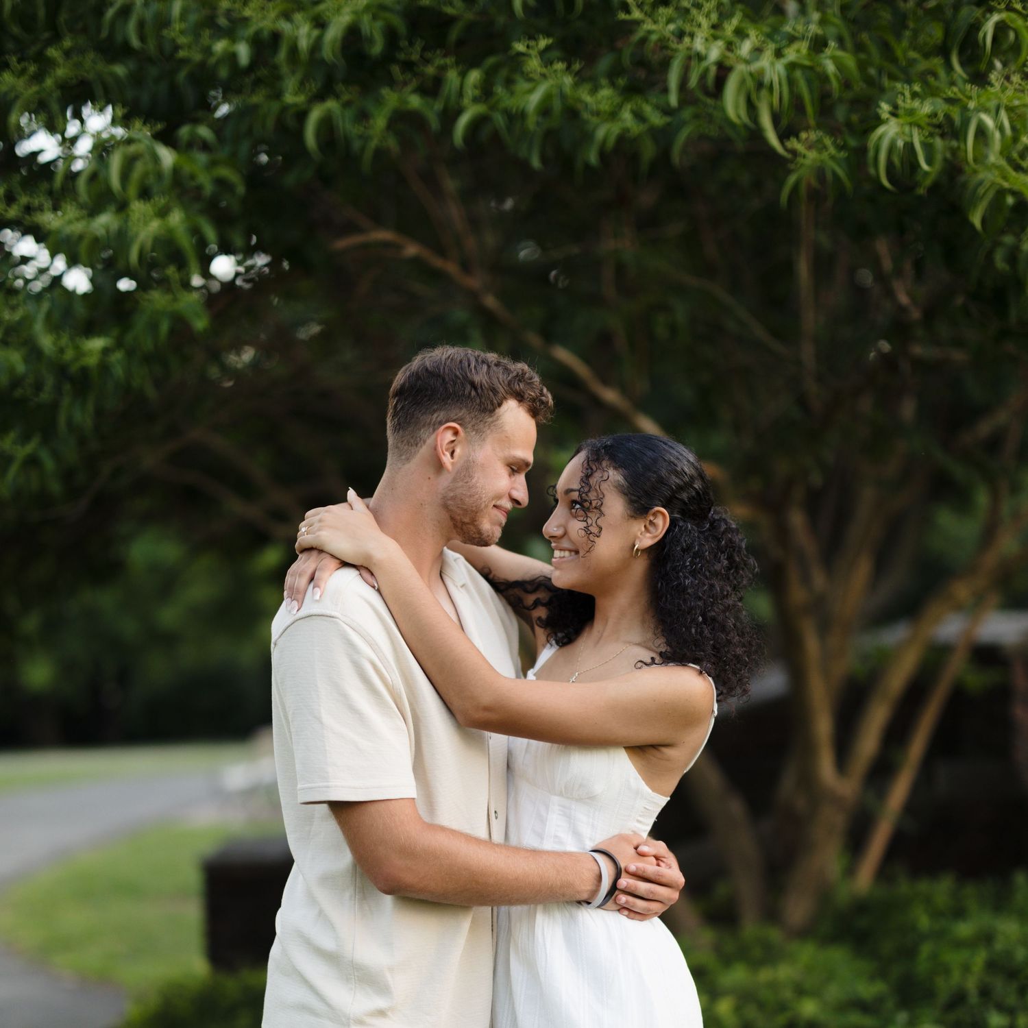 Couple sharing an intimate embrace outdoors on a summer day with lush green trees in the background.