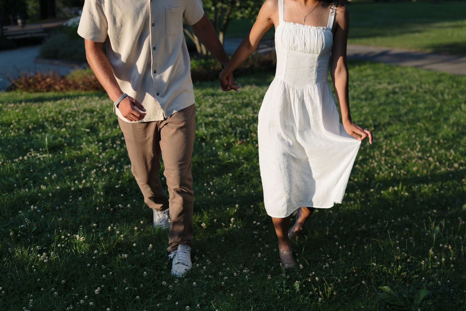 A couple walks together on green grass, with the woman wearing a white summer dress and the man in beige pants.