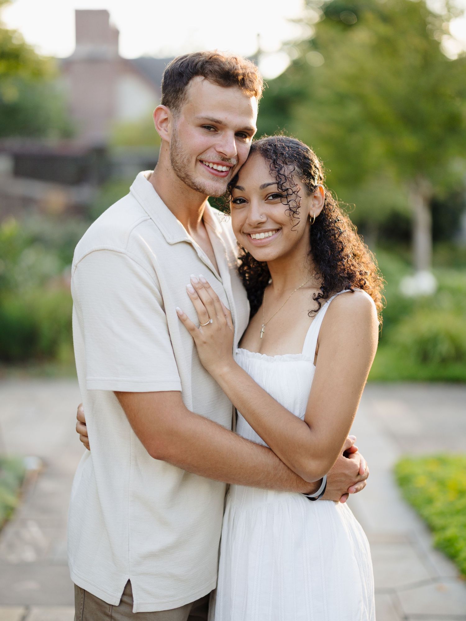 Happy engaged couple in white outfits embrace and smile outdoors in a garden setting during golden hour.