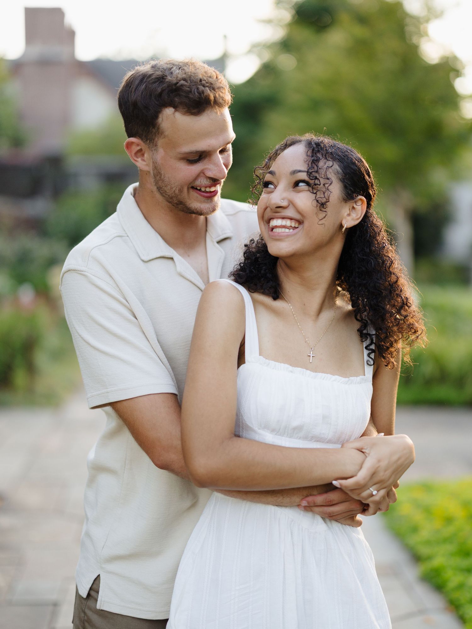 Couple shares a romantic embrace outdoors in white and beige coordinated outfits against a garden background.
