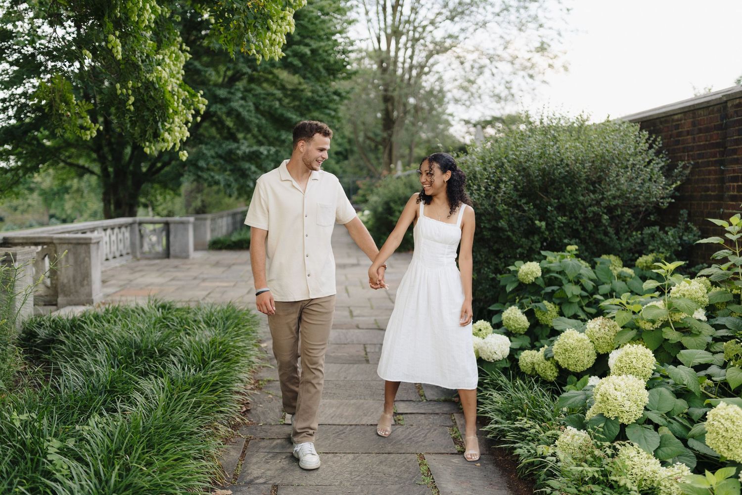 A couple strolls hand in hand along a garden path lined with blooming hydrangeas and lush greenery.