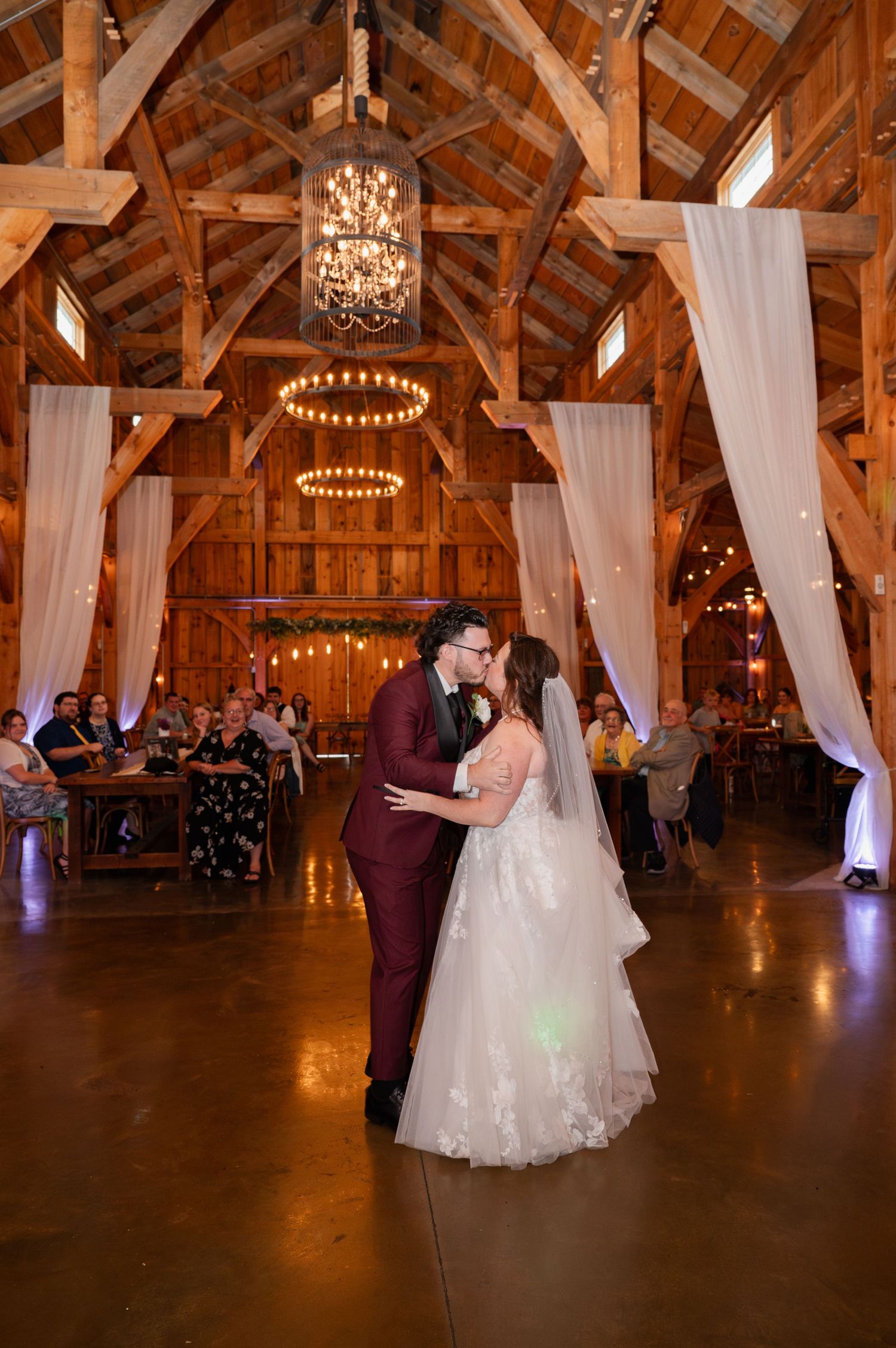 A romantic first dance under warm lighting and chandeliers in an elegant rustic barn venue with flowing white drapes.