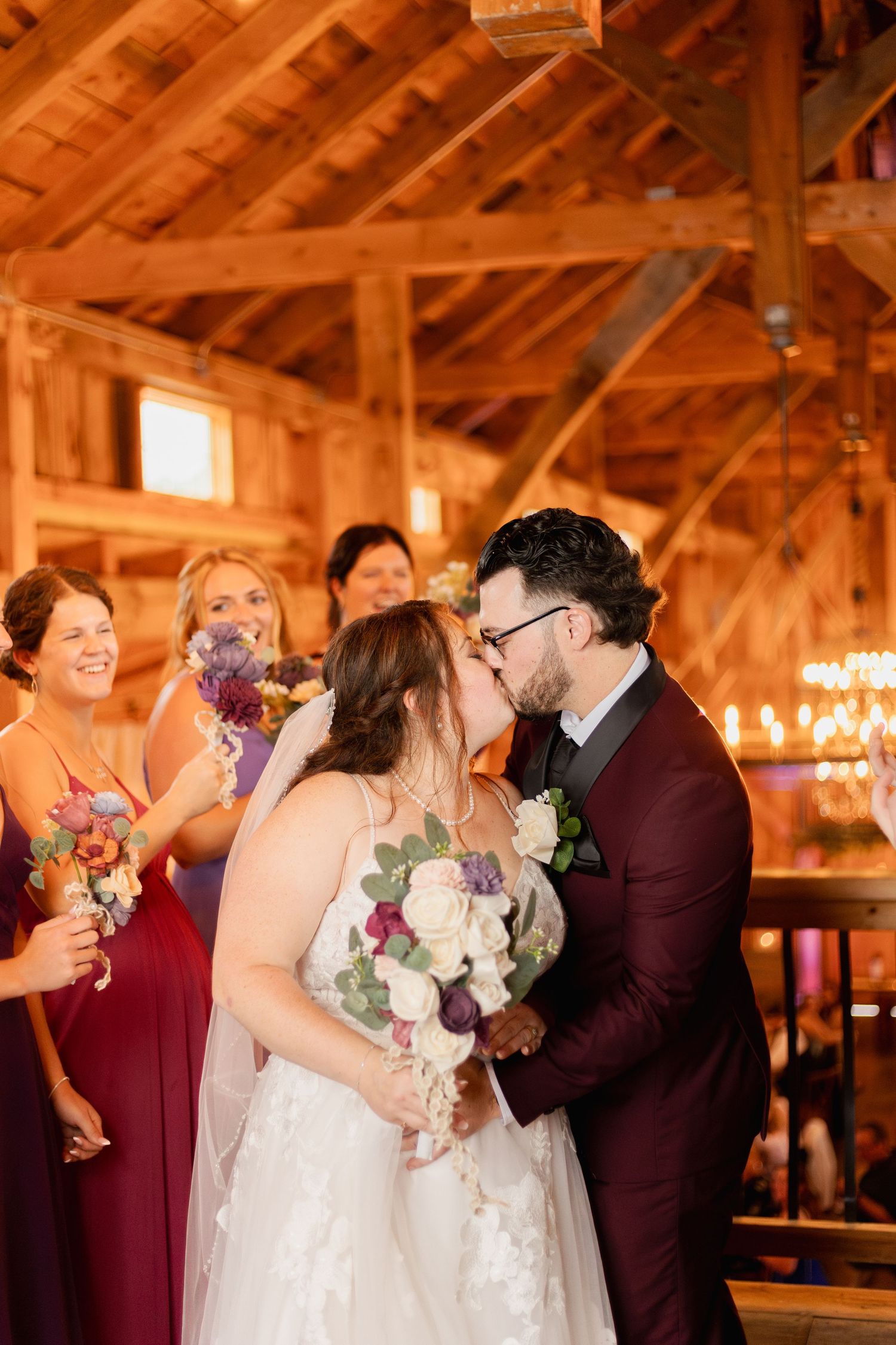 A romantic wedding kiss moment in a rustic wooden barn with burgundy-clad bridesmaids and glowing candlelight.