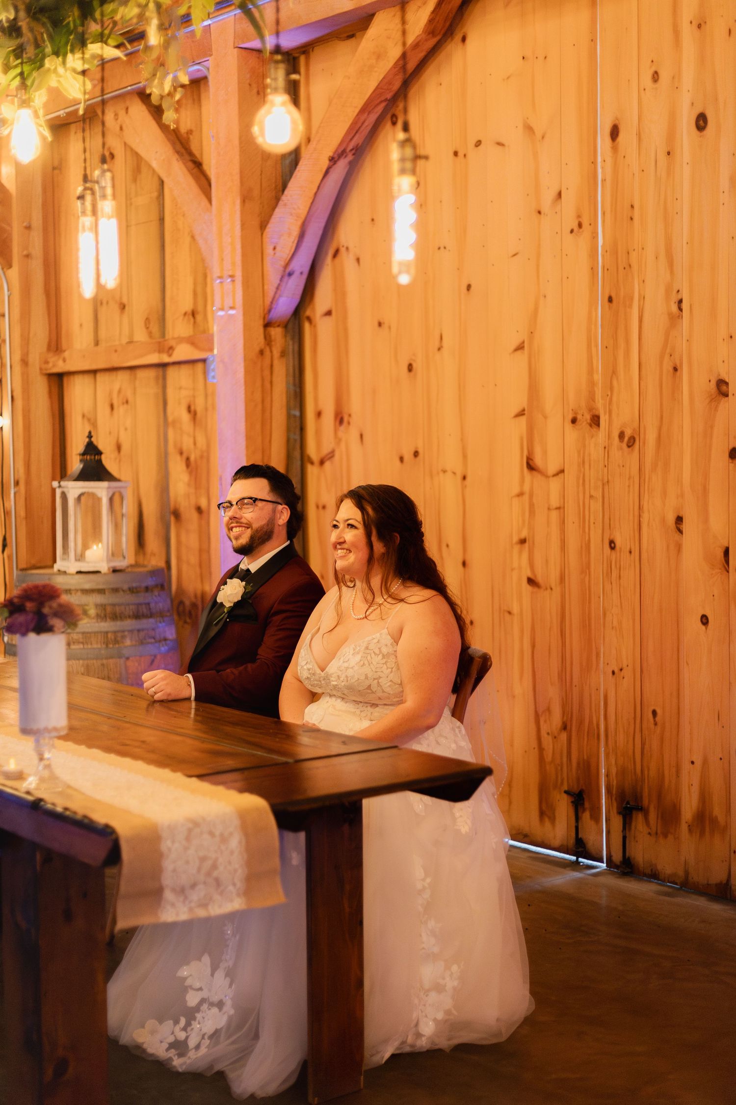 A couple sits together at a rustic wooden table decorated with lights and lanterns in a barn-style venue.