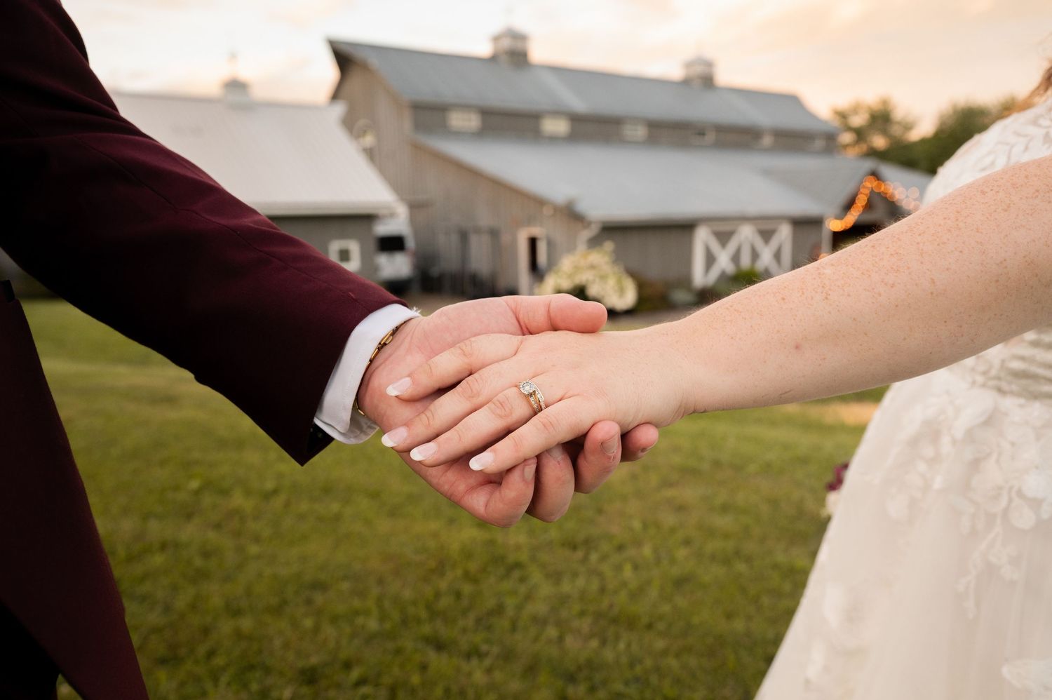 Two hands join together in a tender moment outside a rustic barn wedding venue at sunset.