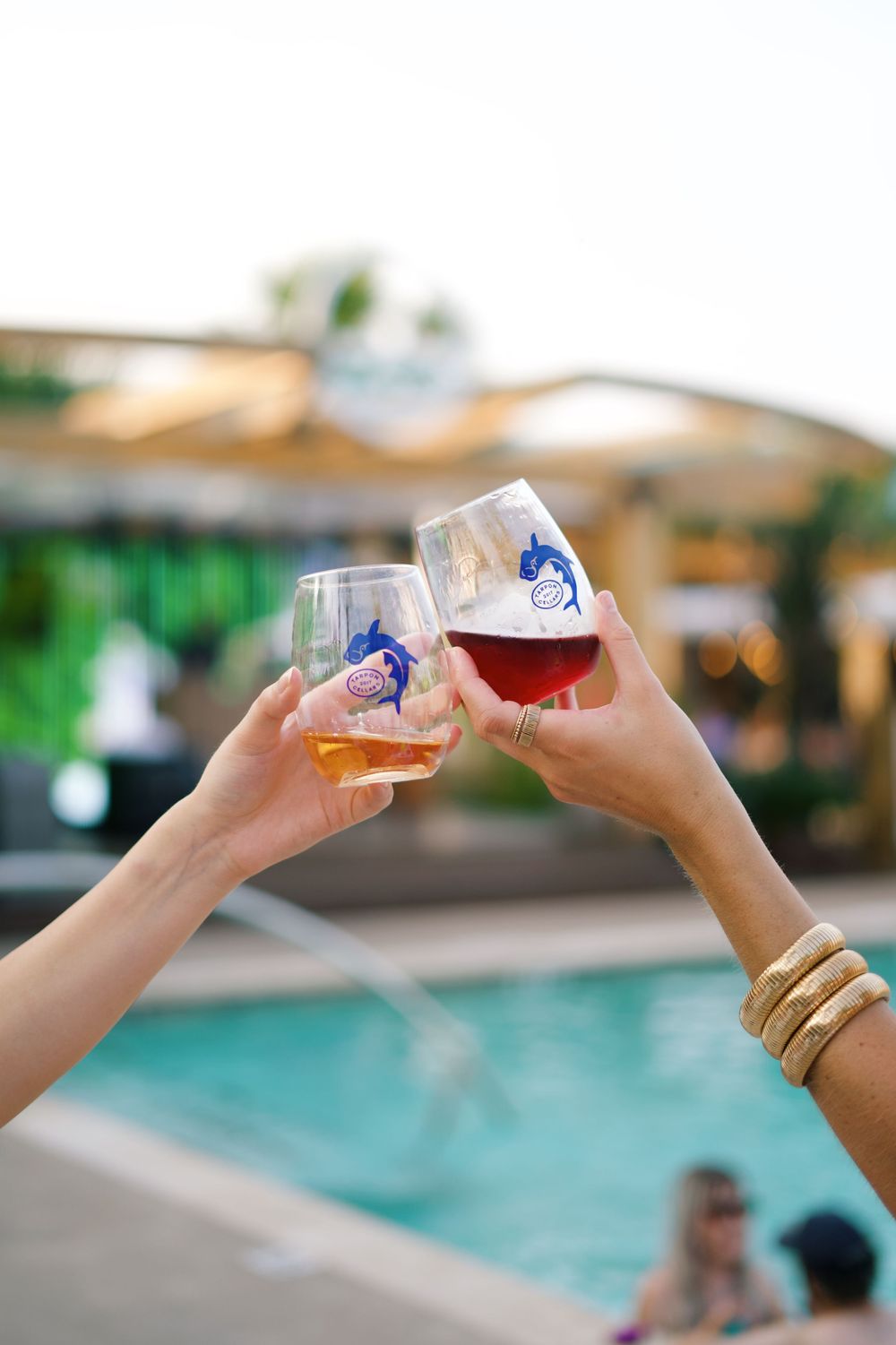 Two hands toasting wine glasses by a swimming pool at sunset.