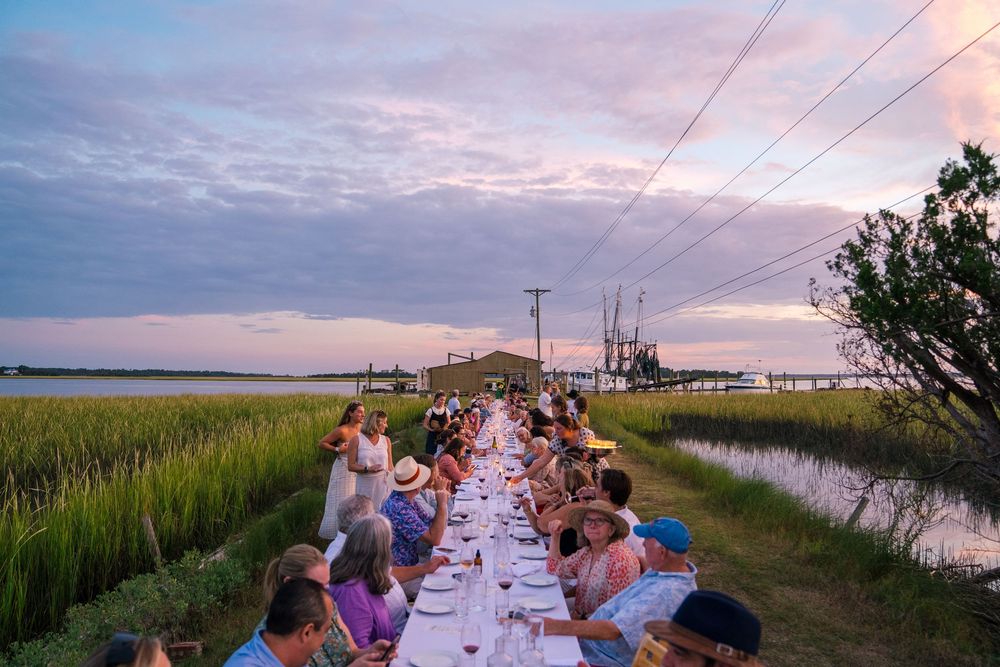 Long outdoor dining table set up along a waterfront at sunset.