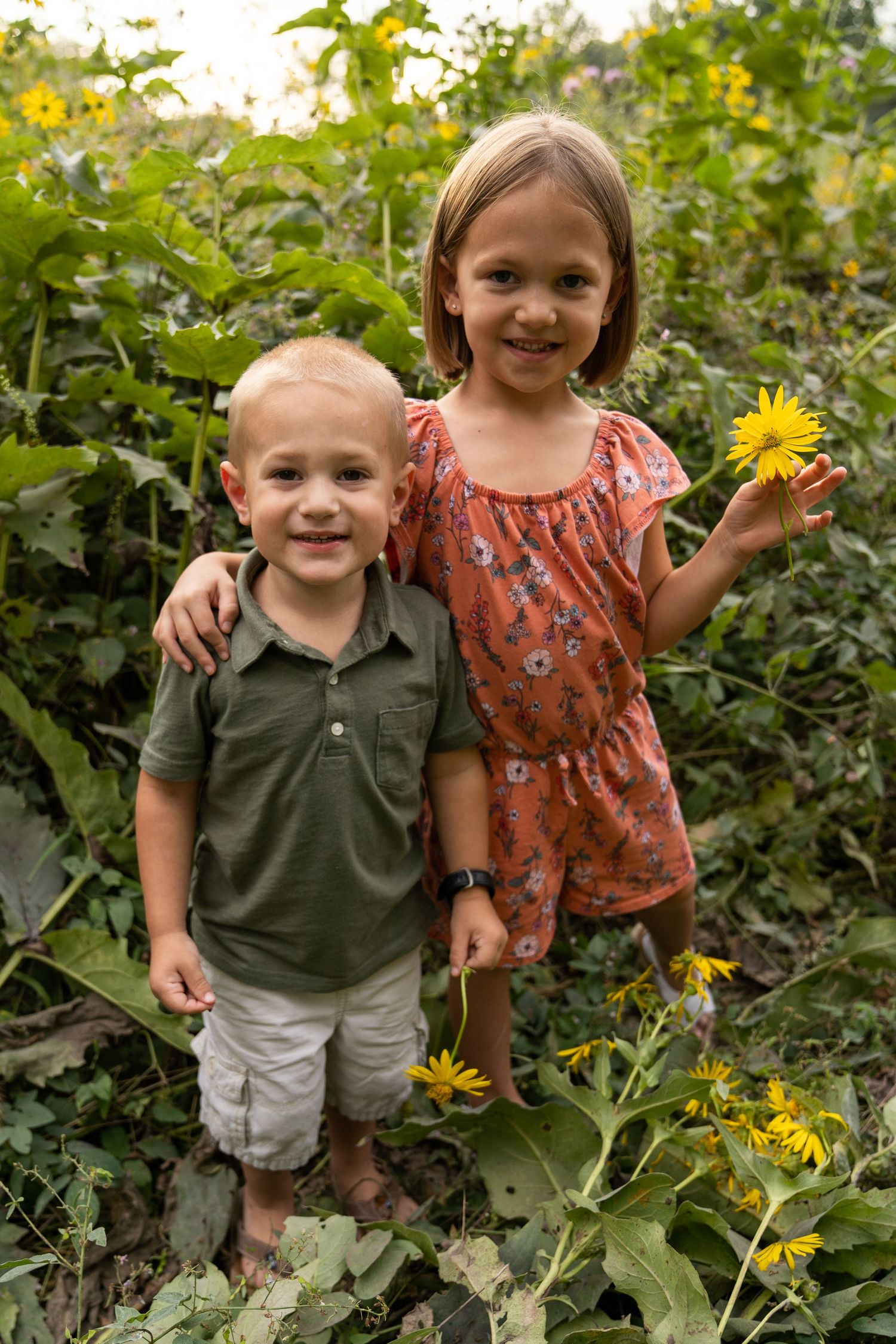 Family portrait of sister and brother standing in a flower patch while sister holds a flower.
