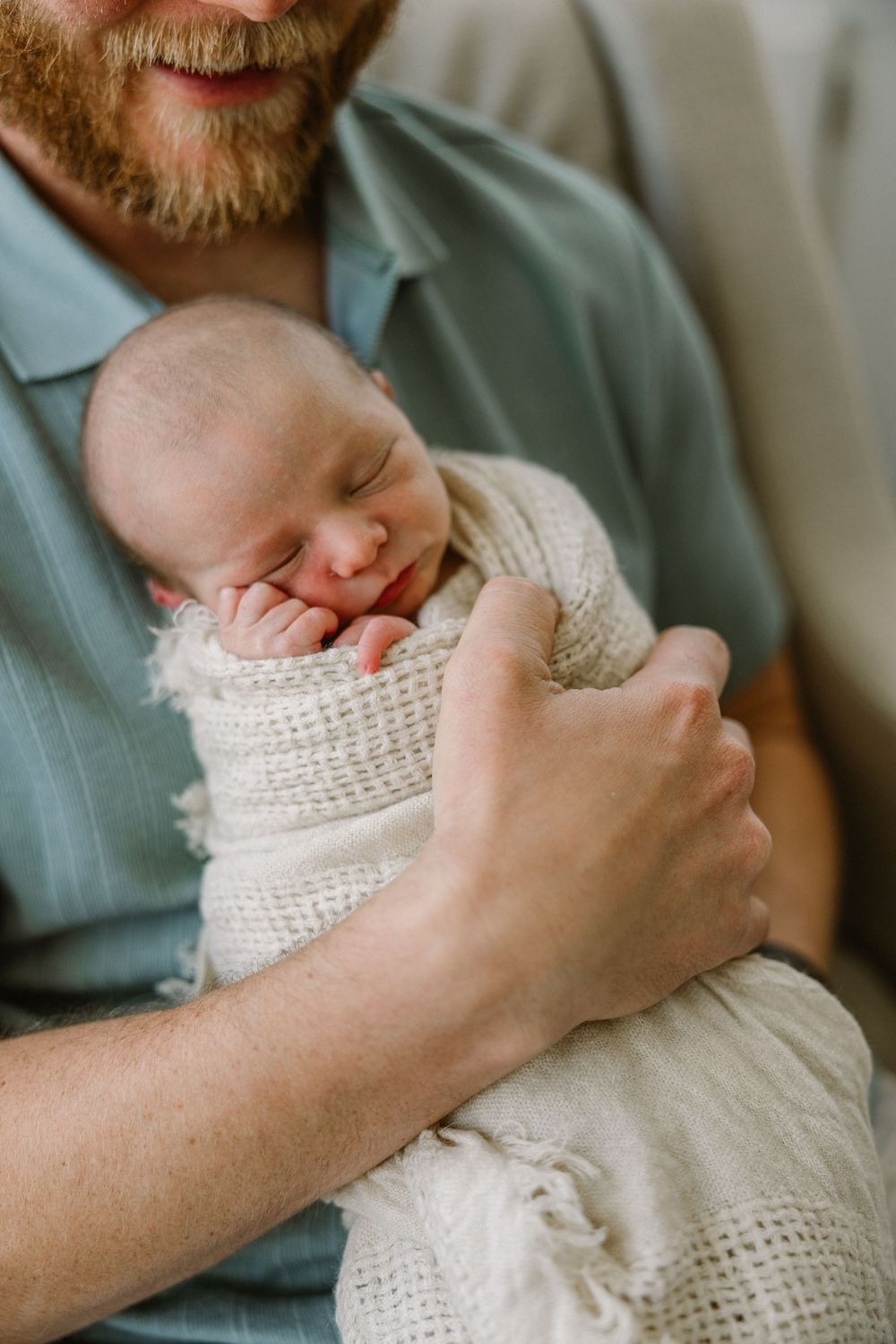 A tender moment shows a sleeping newborn baby swaddled in white knit fabric being cradled in gentle hands.