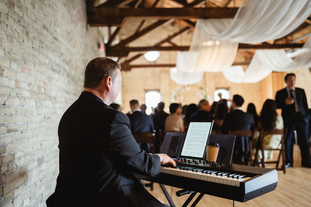 A musician plays keyboard at an elegant indoor venue with exposed brick walls and white drapes hanging from wooden beams.