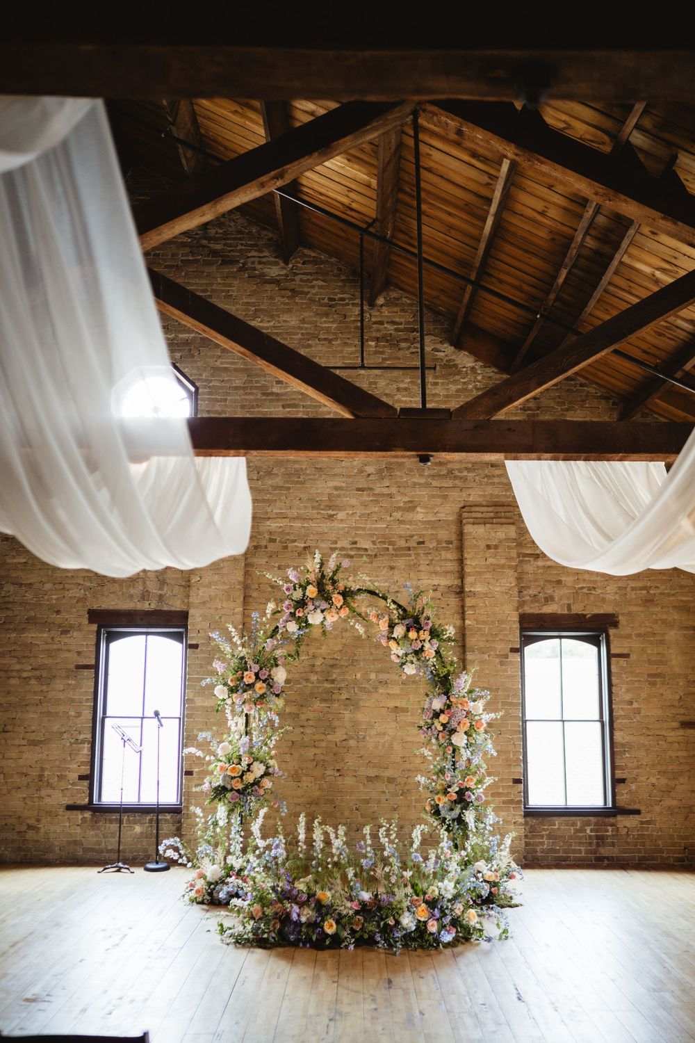 A wedding arch decorated with flowers and greenery stands in a rustic brick venue with exposed wooden beams and flowy drapes.