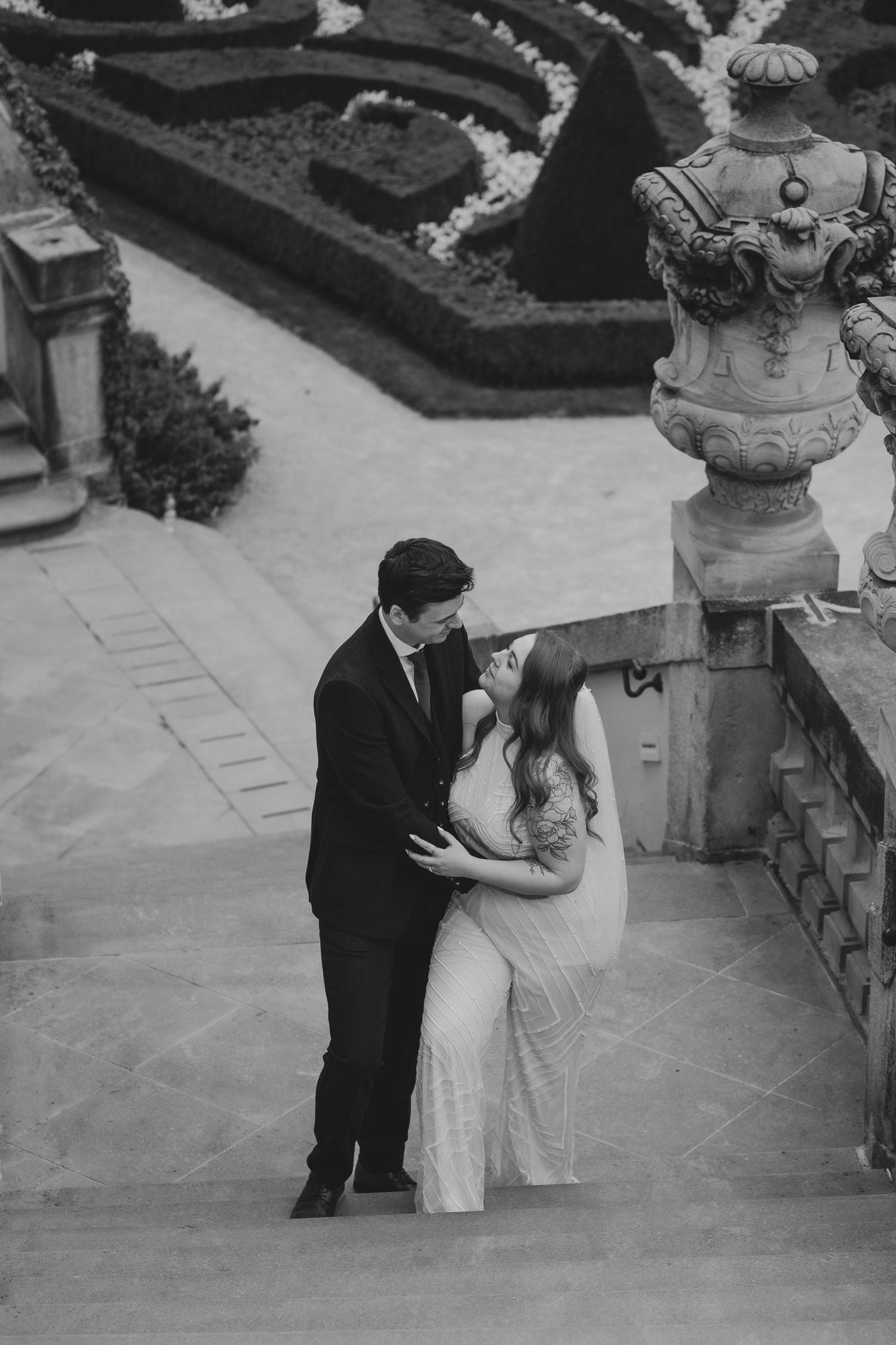 A black and white photo of an intimate couple's moment on ornate stone steps next to a decorative garden urn.