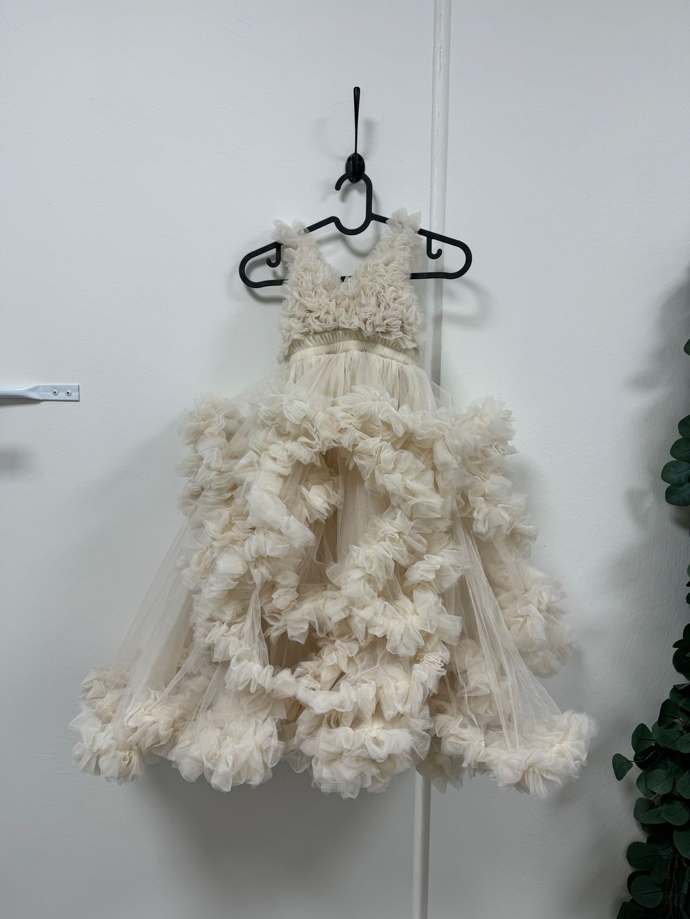 An ornate cream-colored ruffled tulle dress with lace bodice and tiered skirts hangs on a black hanger against white wall.