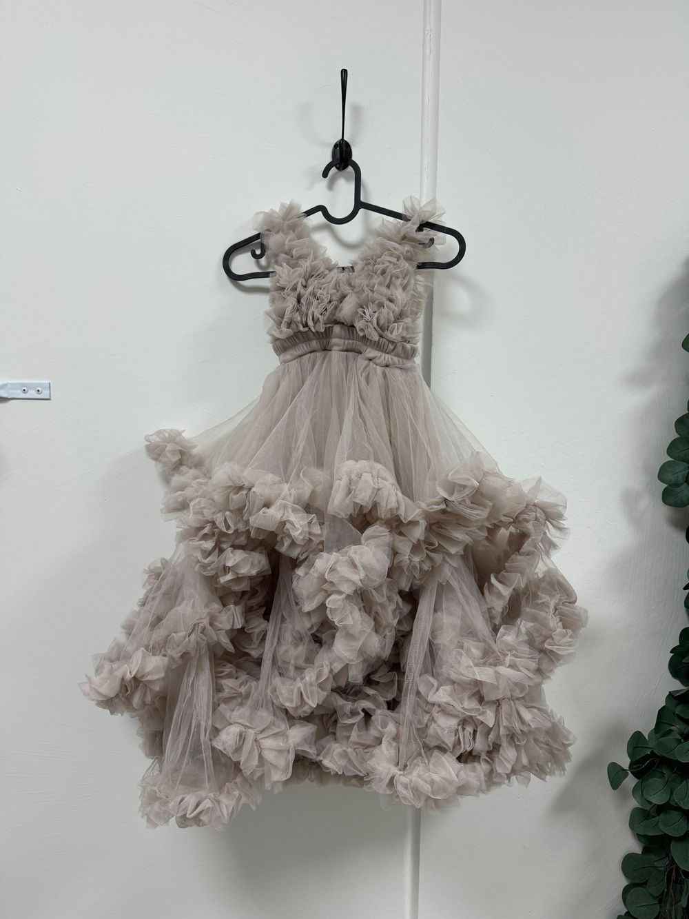 A frilly beige tulle dress with ruffled layers and embellished bodice hangs on a black hanger against a white wall.
