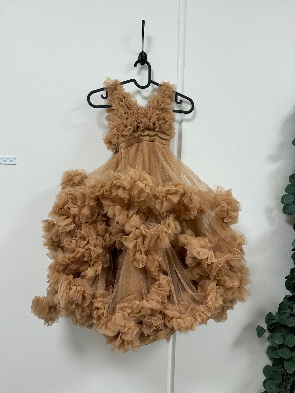 A luxurious caramel-colored tulle dress with ruffled layers and bodice hangs on a black hanger against a white wall.