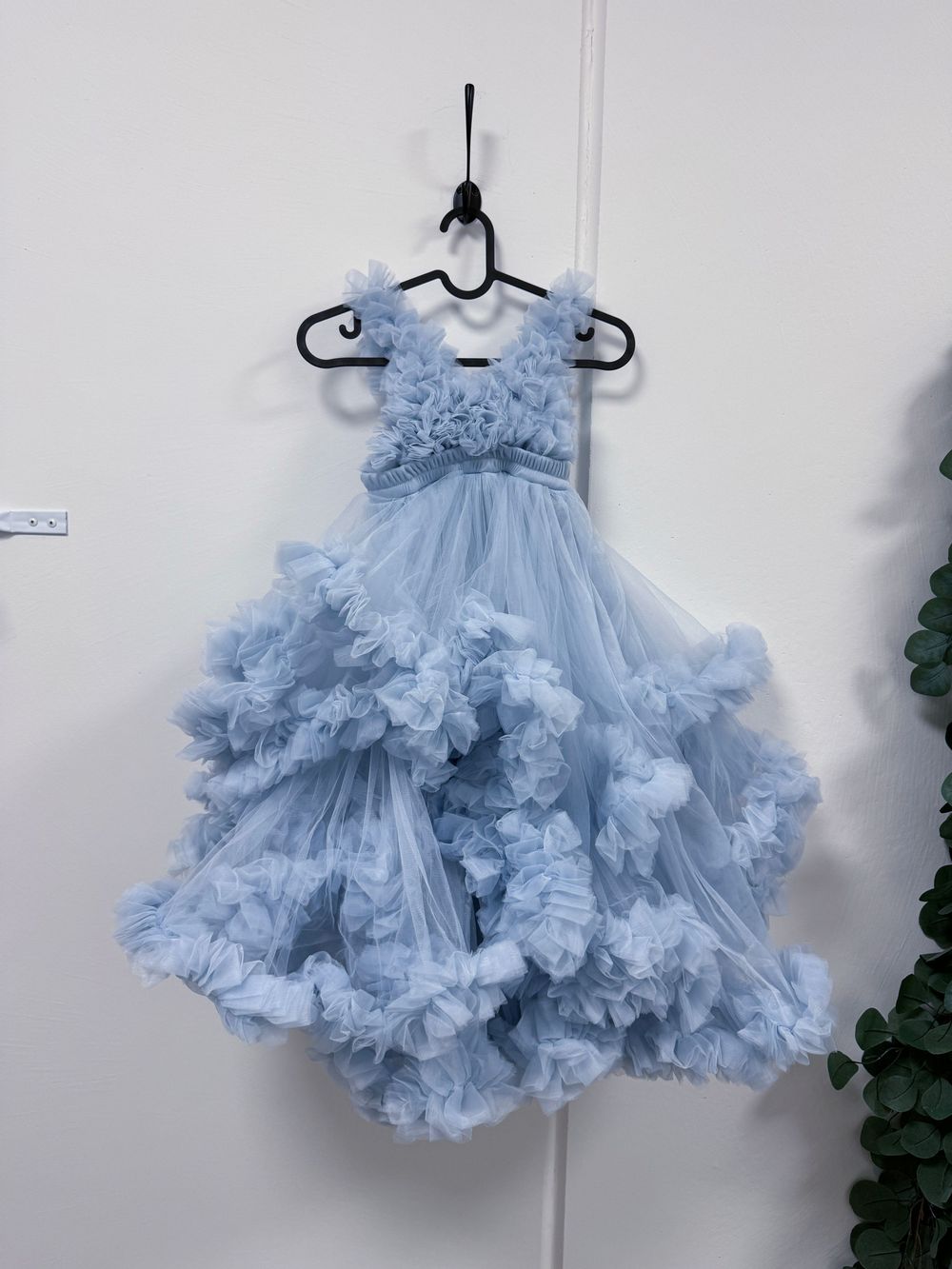 A delicate light blue tulle dress with ruffled layers and floral accents hangs on a white hanger against a white wall.