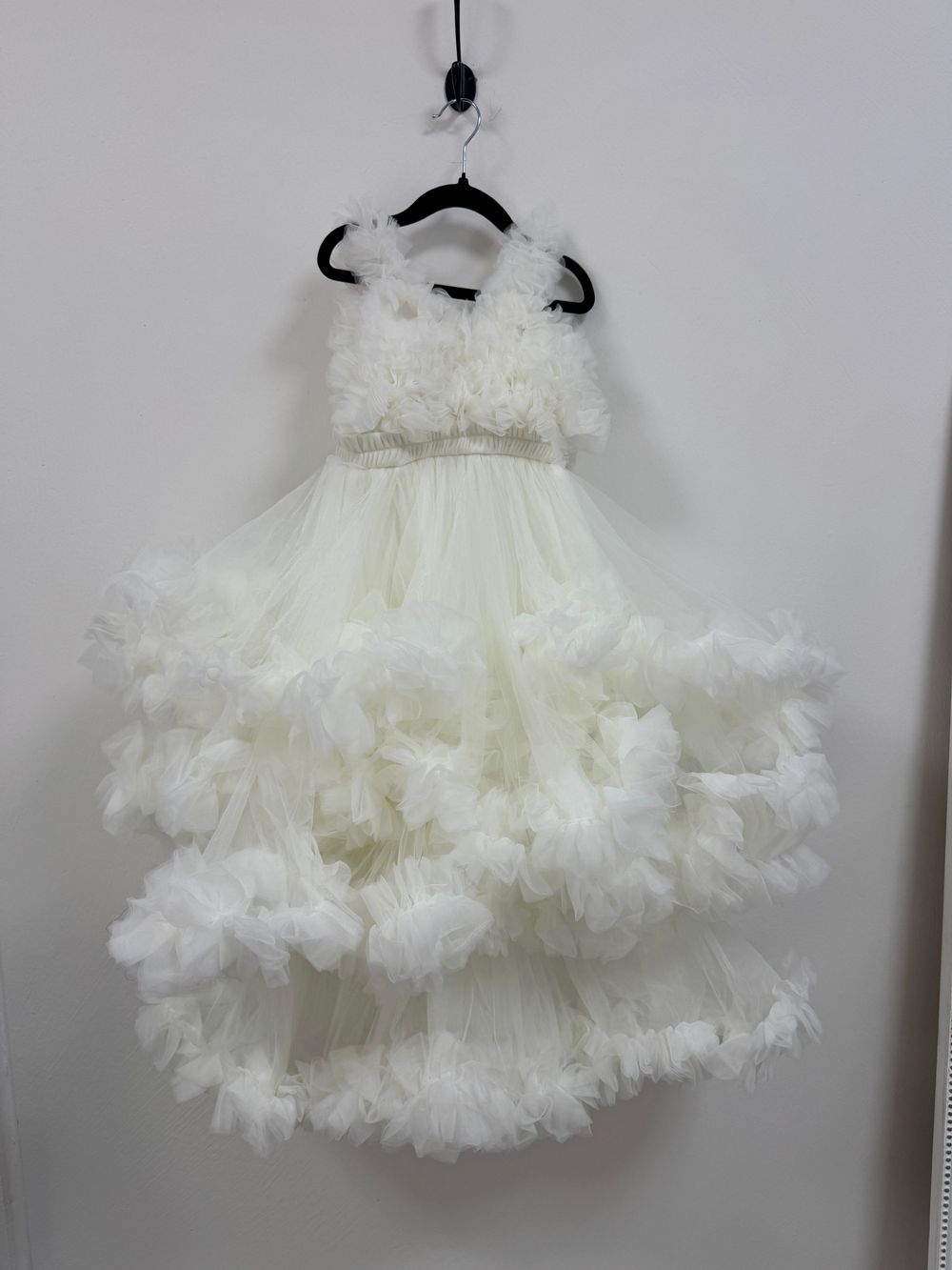 A collection of white puffy tulle petticoat dresses with ruffled layers hanging on display against a gray wall.